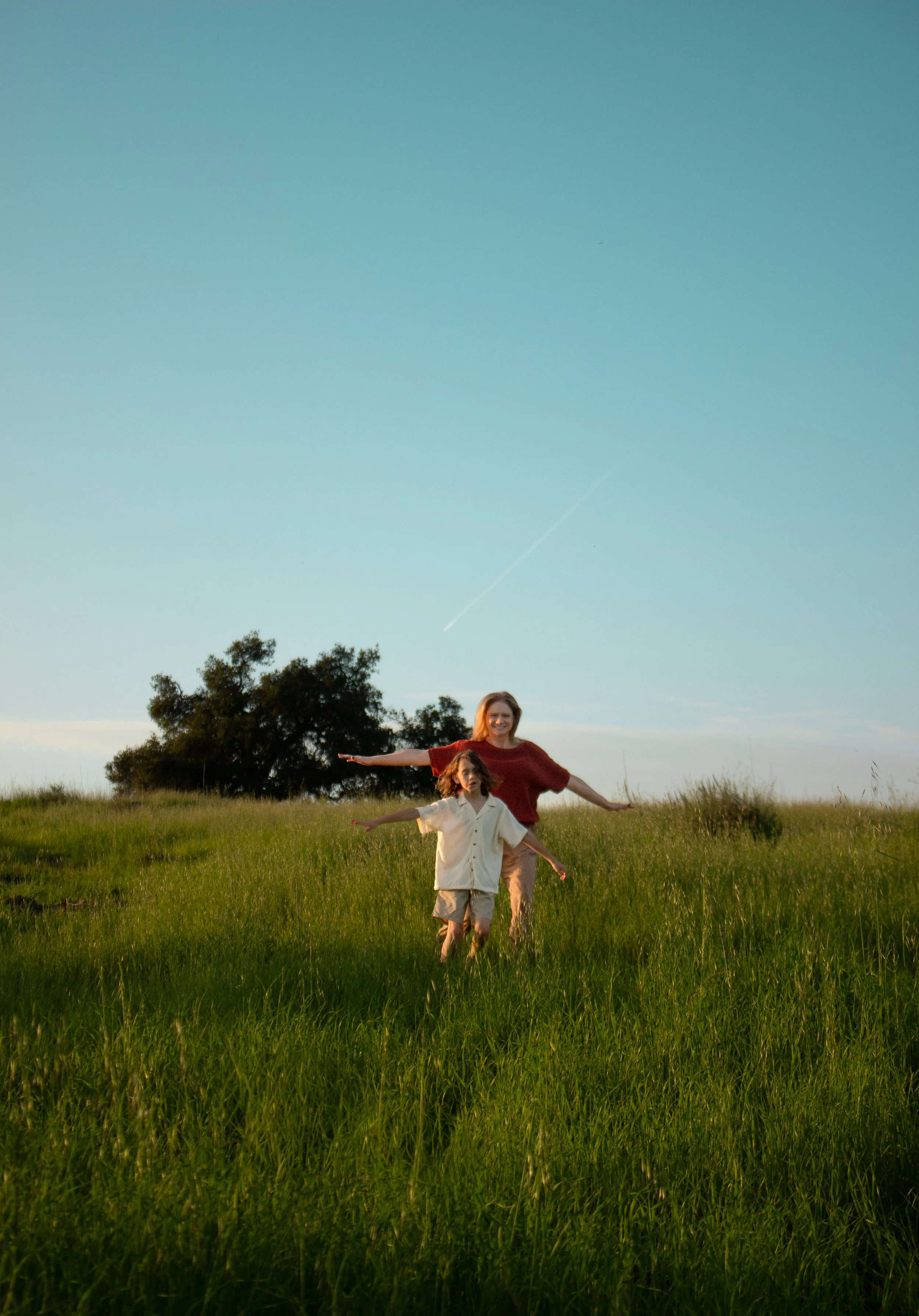 A woman and a child playing in a grassy field during sunset with a tree in the background.