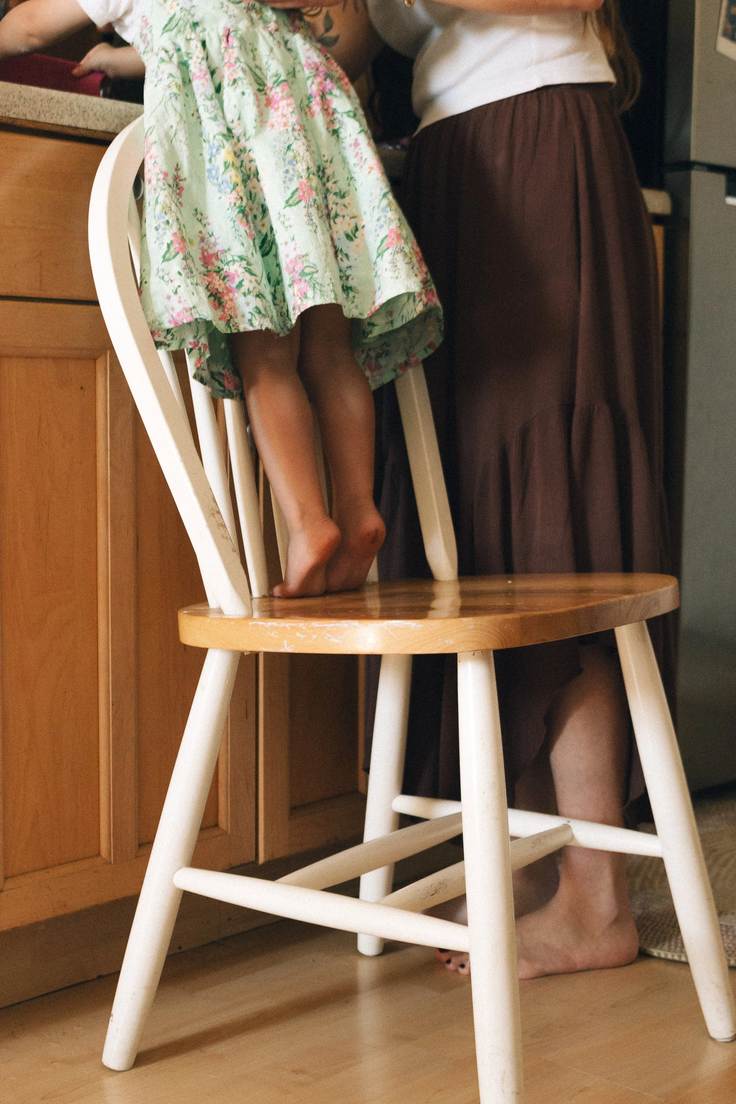 Child standing on a wooden chair with feet on the seat, wearing a floral dress, with an adult nearby in a kitchen setting.