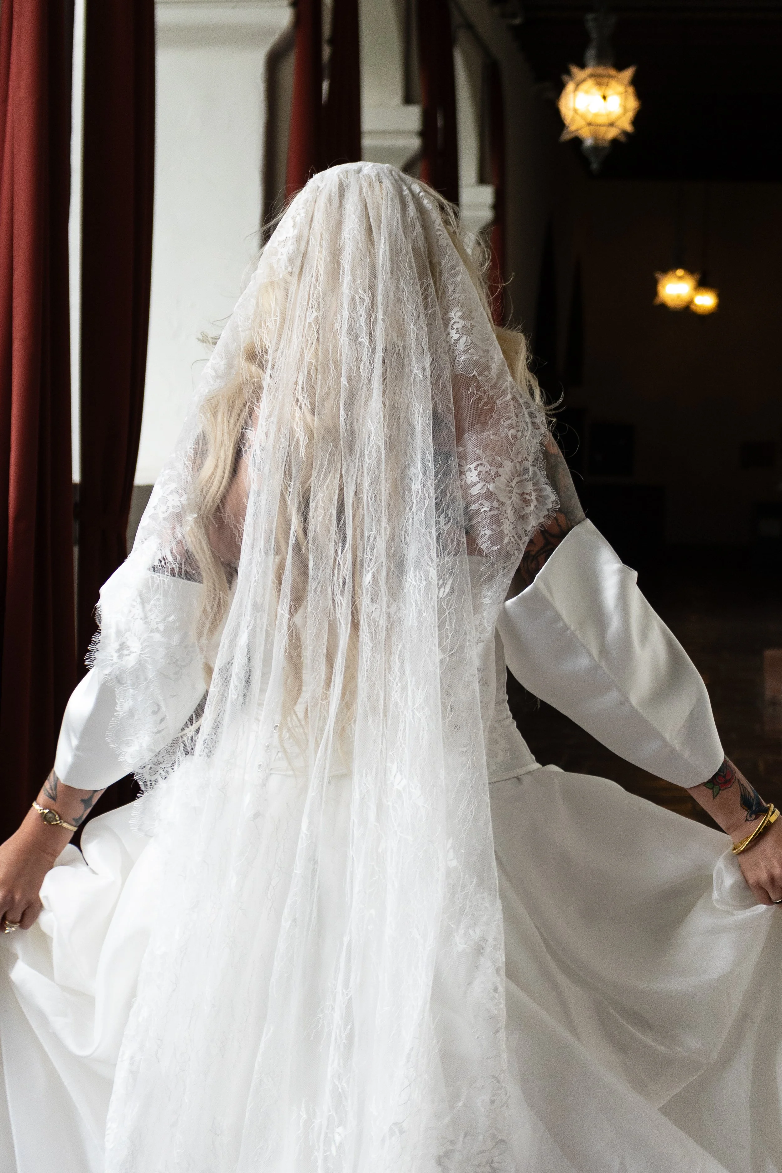 A bride wearing a white wedding dress and lace veil, adjusting her gown in a dimly lit room with hanging lamps and dark red curtains at a Santa Barbara wedding venue