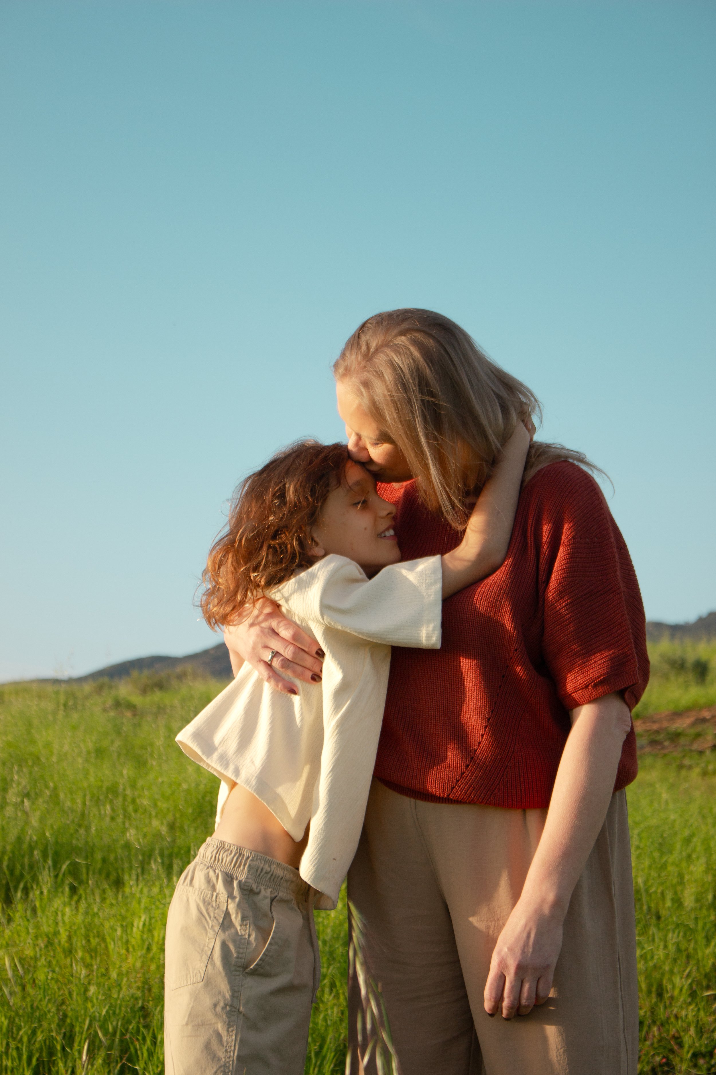 A woman and her son sharing a hug in a grassy outdoor setting under a clear blue sky.