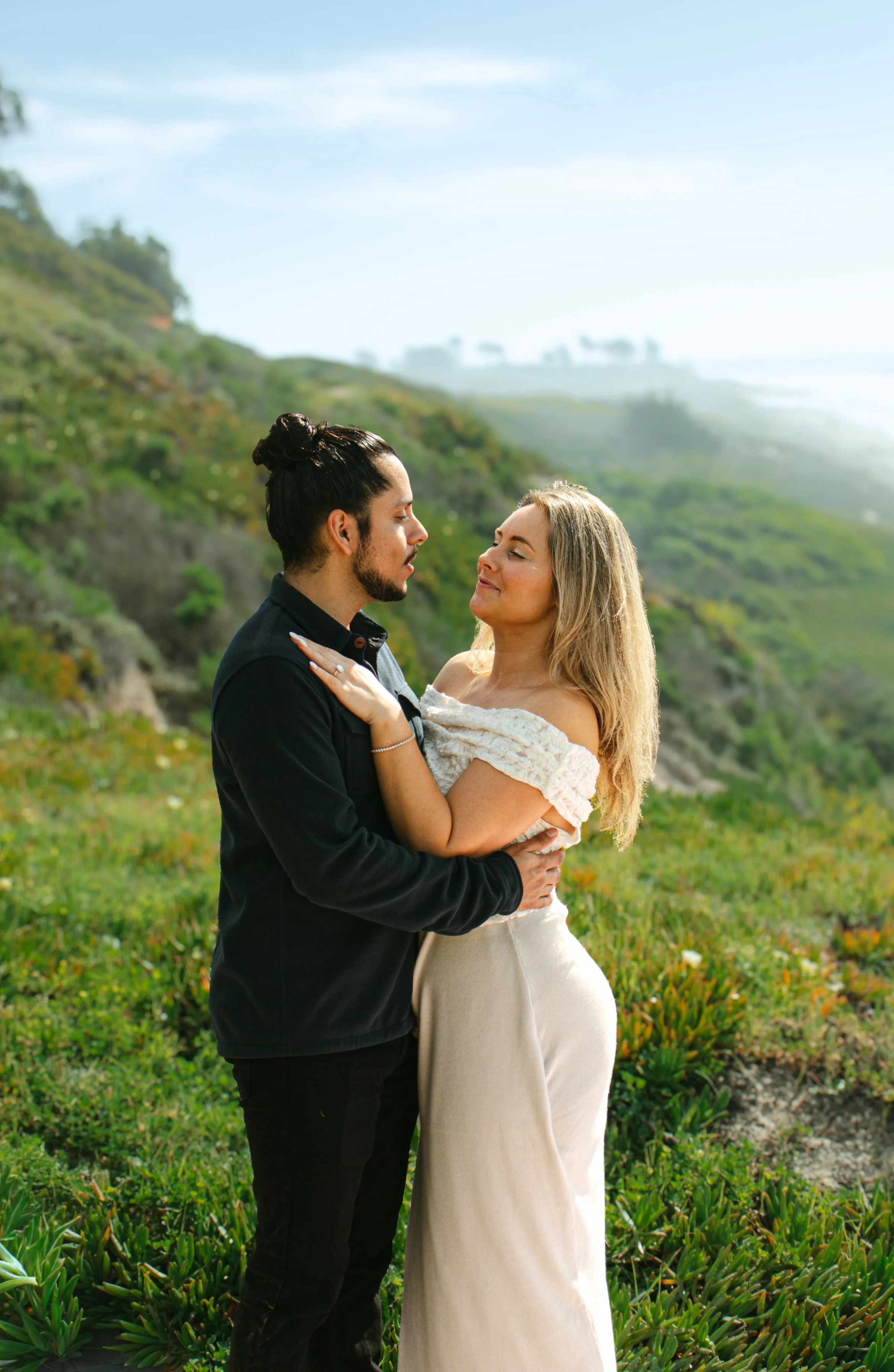 A couple standing outdoors in a grassy area, embracing and looking at each other with a hillside and cloudy sky in the background after getting engaged in Santa Barbara. 