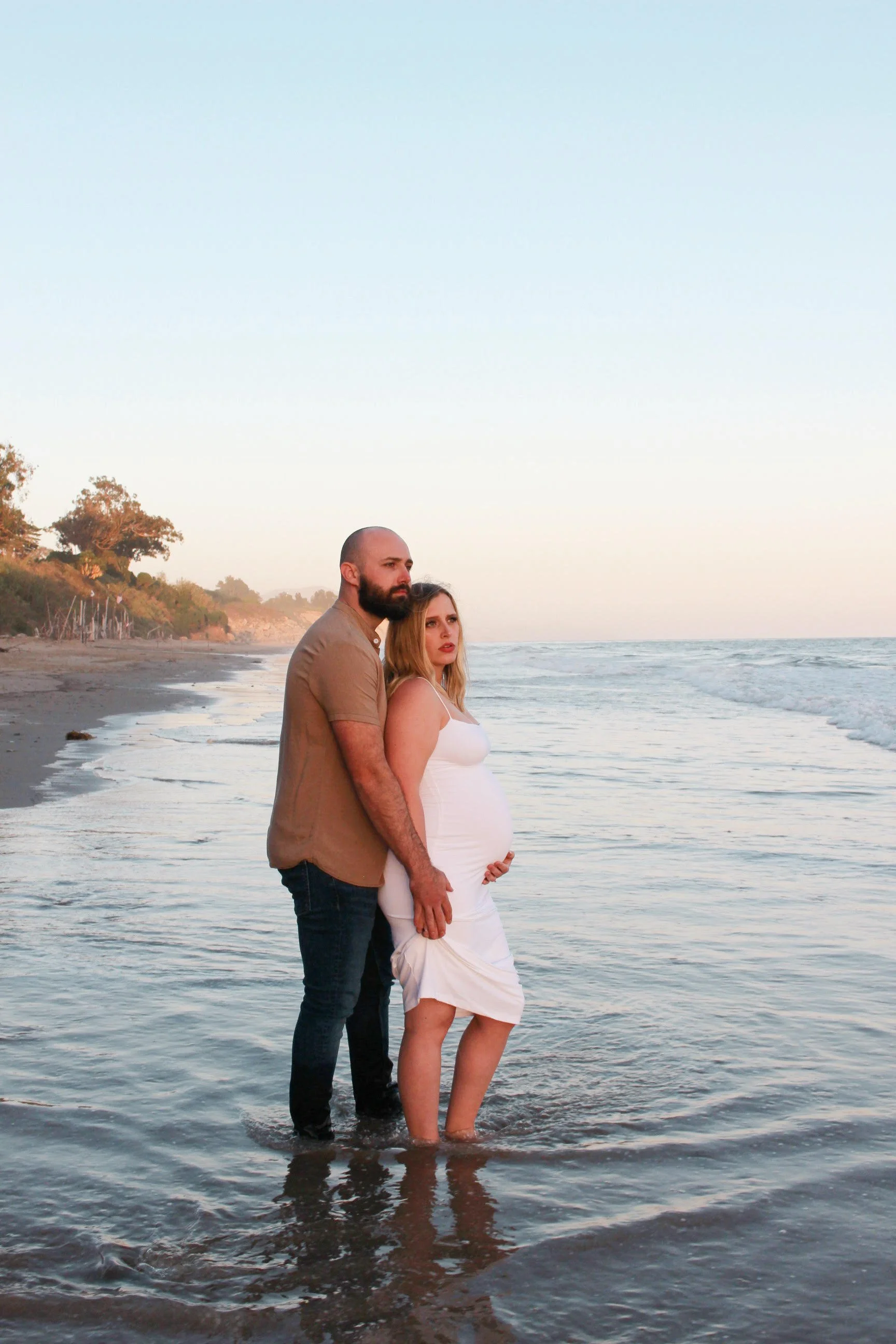 A pregnant woman and her husband hold each other and stand in the water at the beach during sunset in Carpenteria 