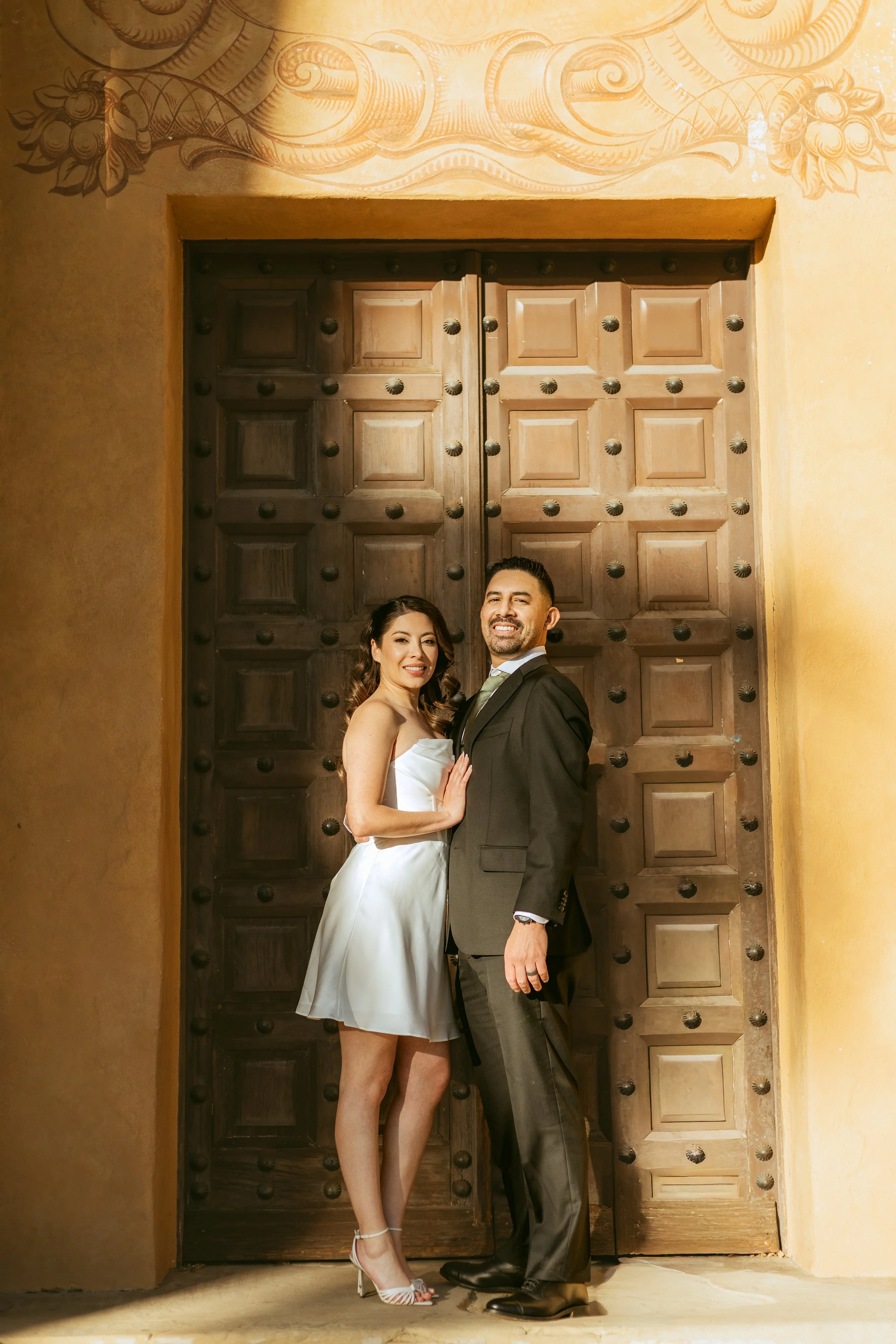 A couple dressed in formal attire, standing in front of large wooden doors, with the woman in a white dress and heels and the man in a black suit, smiling.