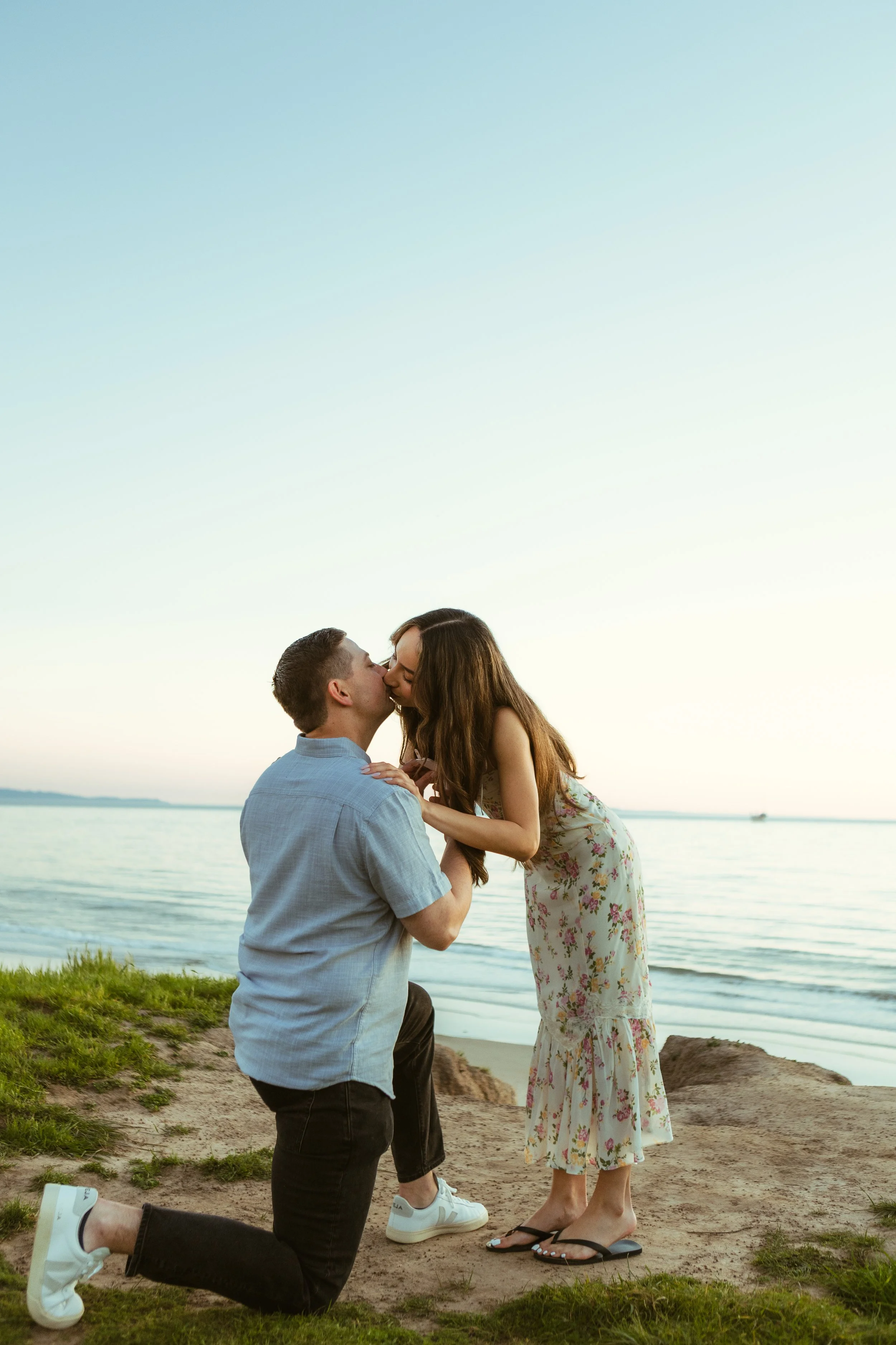 A man is proposing to a woman on the beach at sunset. The man is on one knee, holding the woman's hand, while the woman is leaning in for a kiss or to accept the proposal. The background shows the ocean and a clear sky.