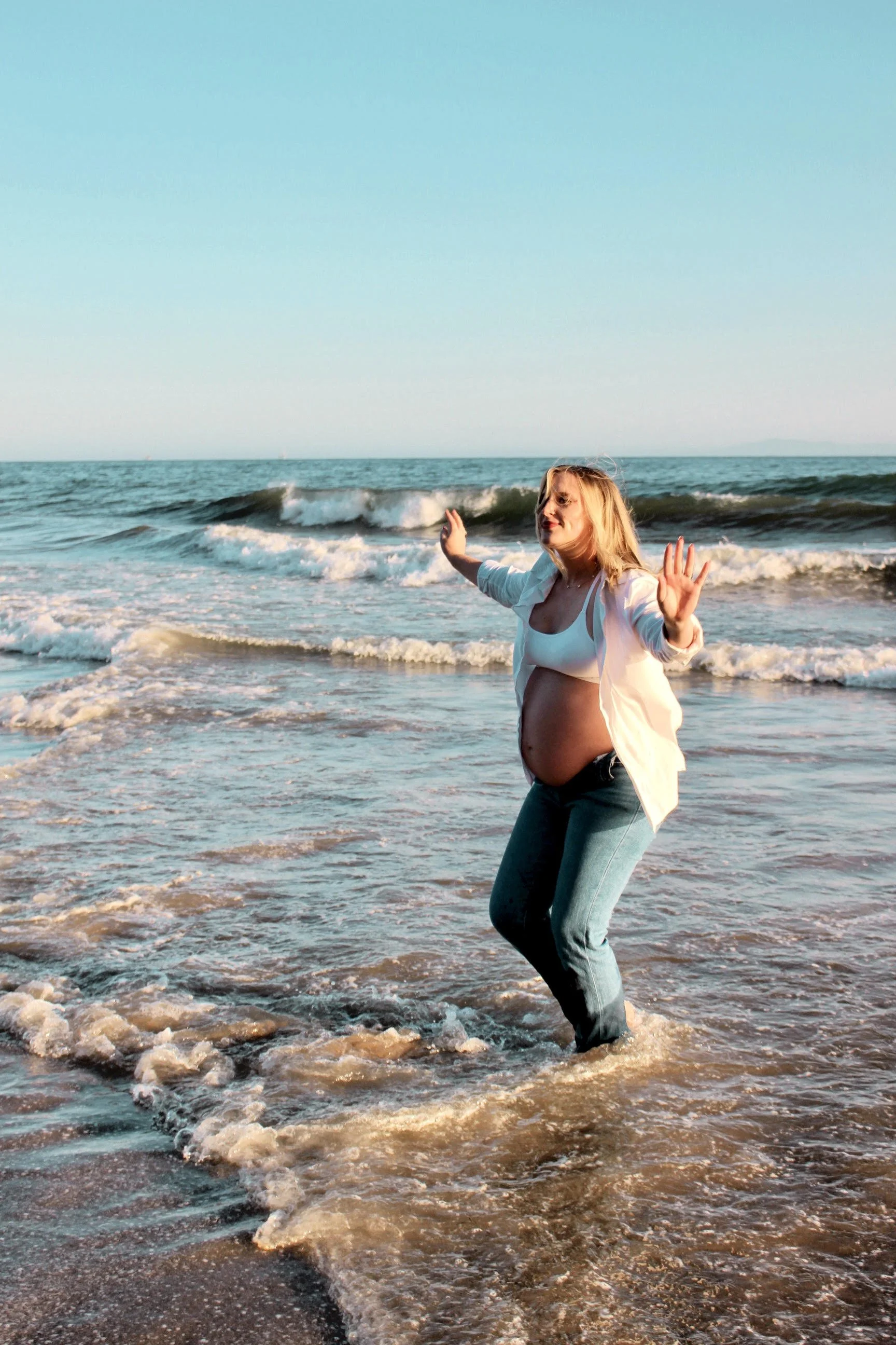 Pregnant woman standing in ocean waves at sunset, raising her hands with a smile.