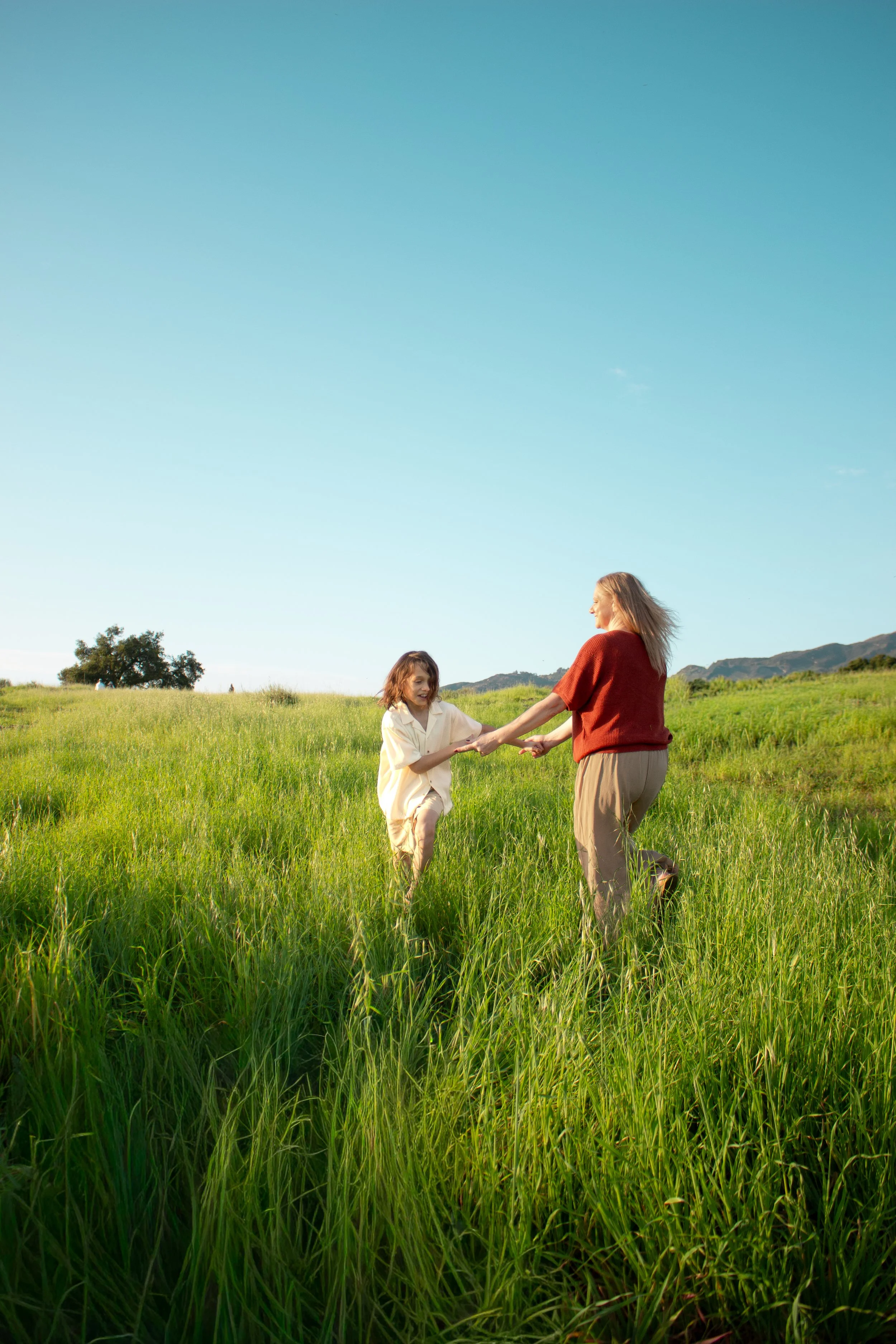 A woman and a young boy playing and holding hands in a lush green field under a clear blue sky.
