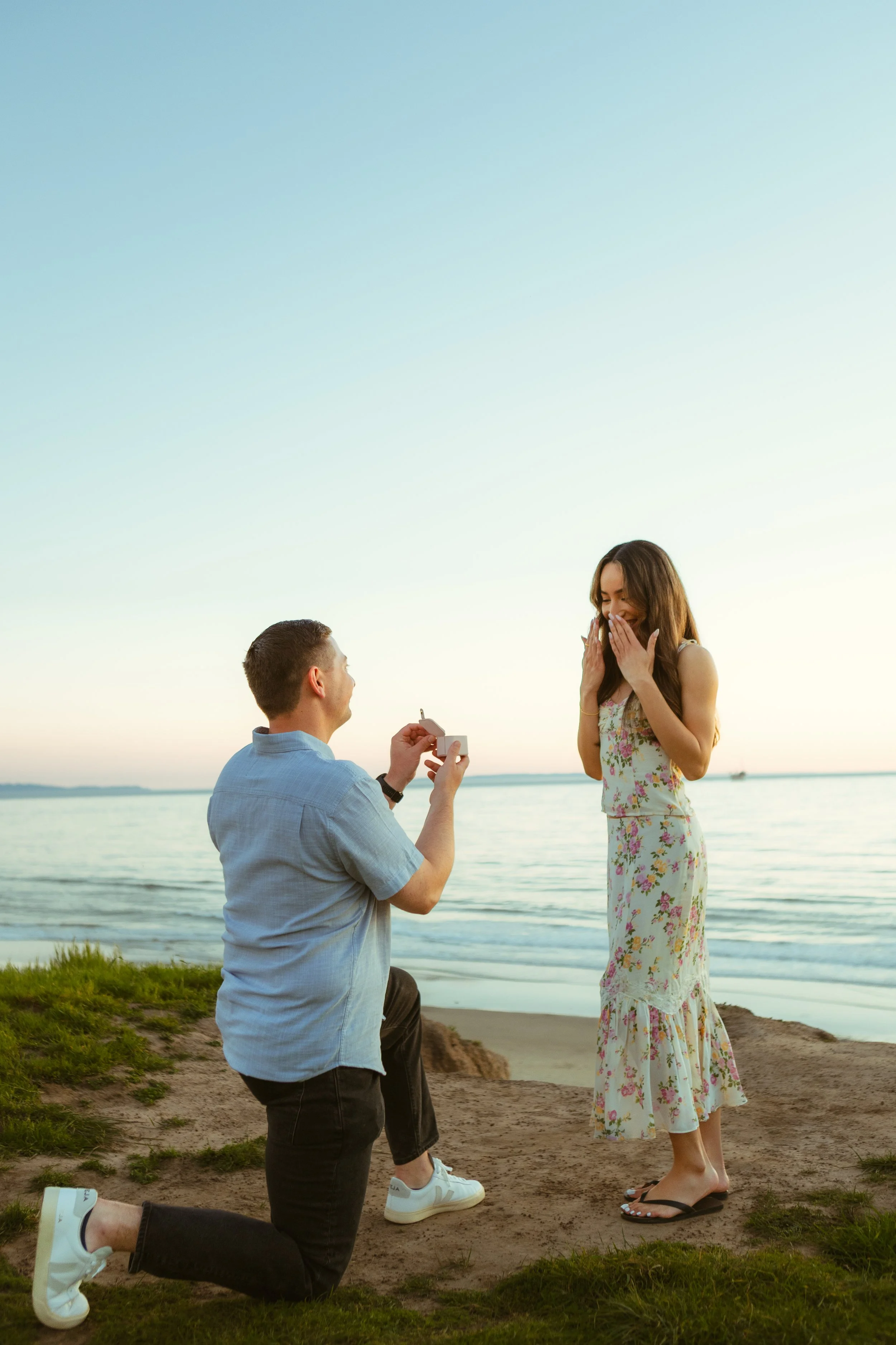 A man proposing marriage to a woman on the beach during sunset, with the man kneeling and holding out a ring box, and the woman smiling with surprise, covering her mouth with her hands.
