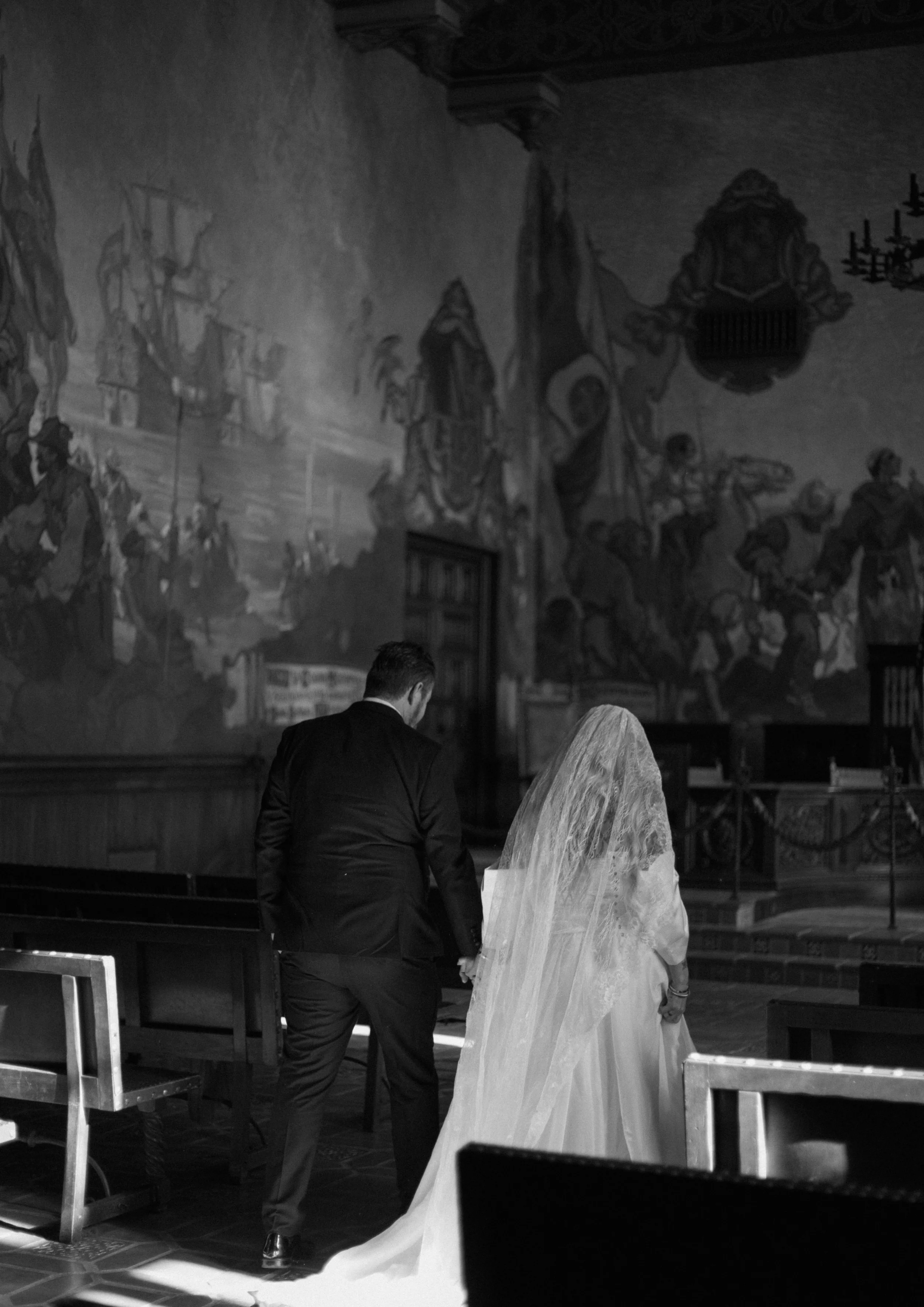 A bride and groom holding hands, walking inside a church with a mural featuring ships and historical scenes in Santa Barbara