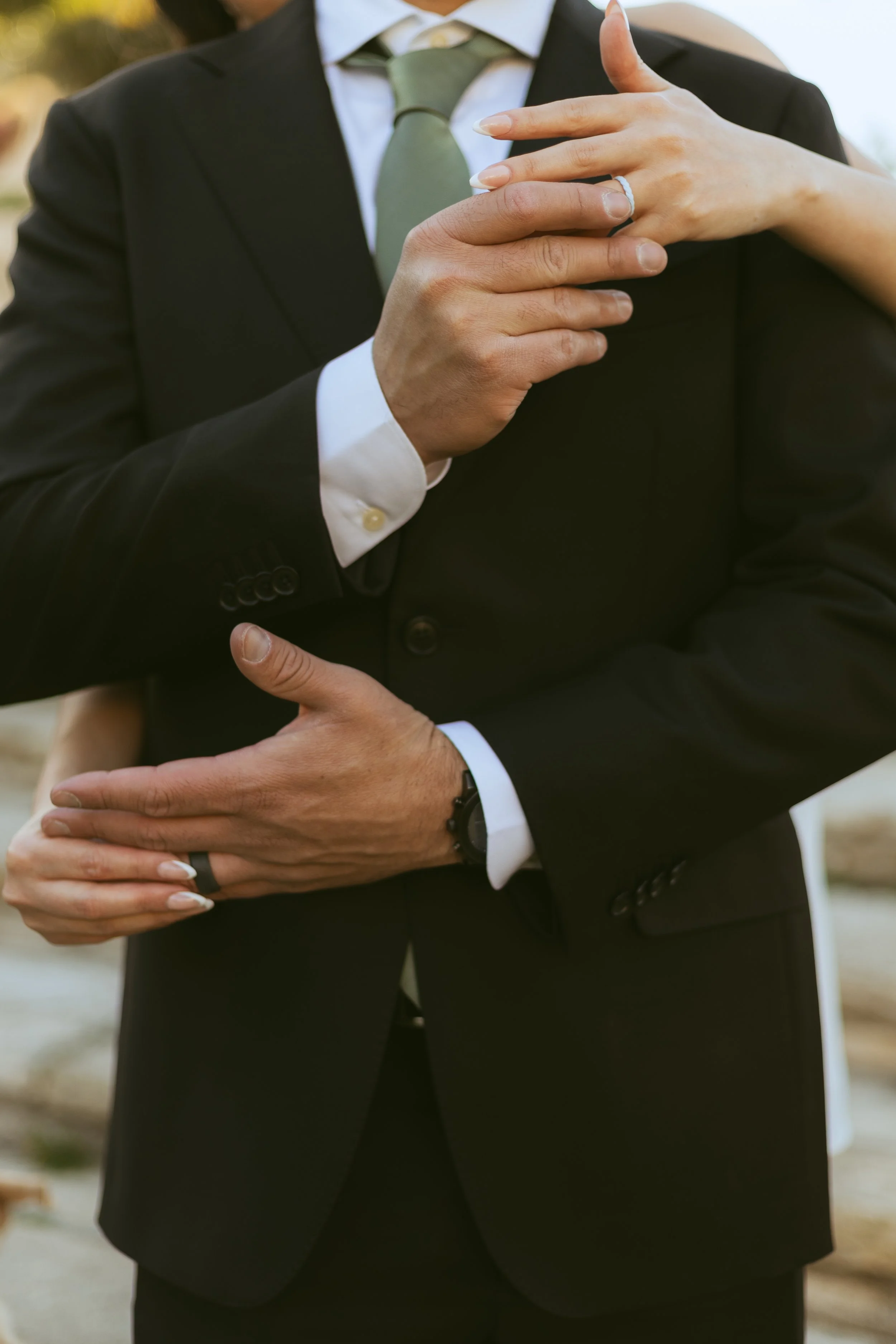 Close-up of a couple dancing, showing the man in a black suit with a white shirt and green tie, and a woman with a painted manicure, wearing a ring, embracing outdoors.