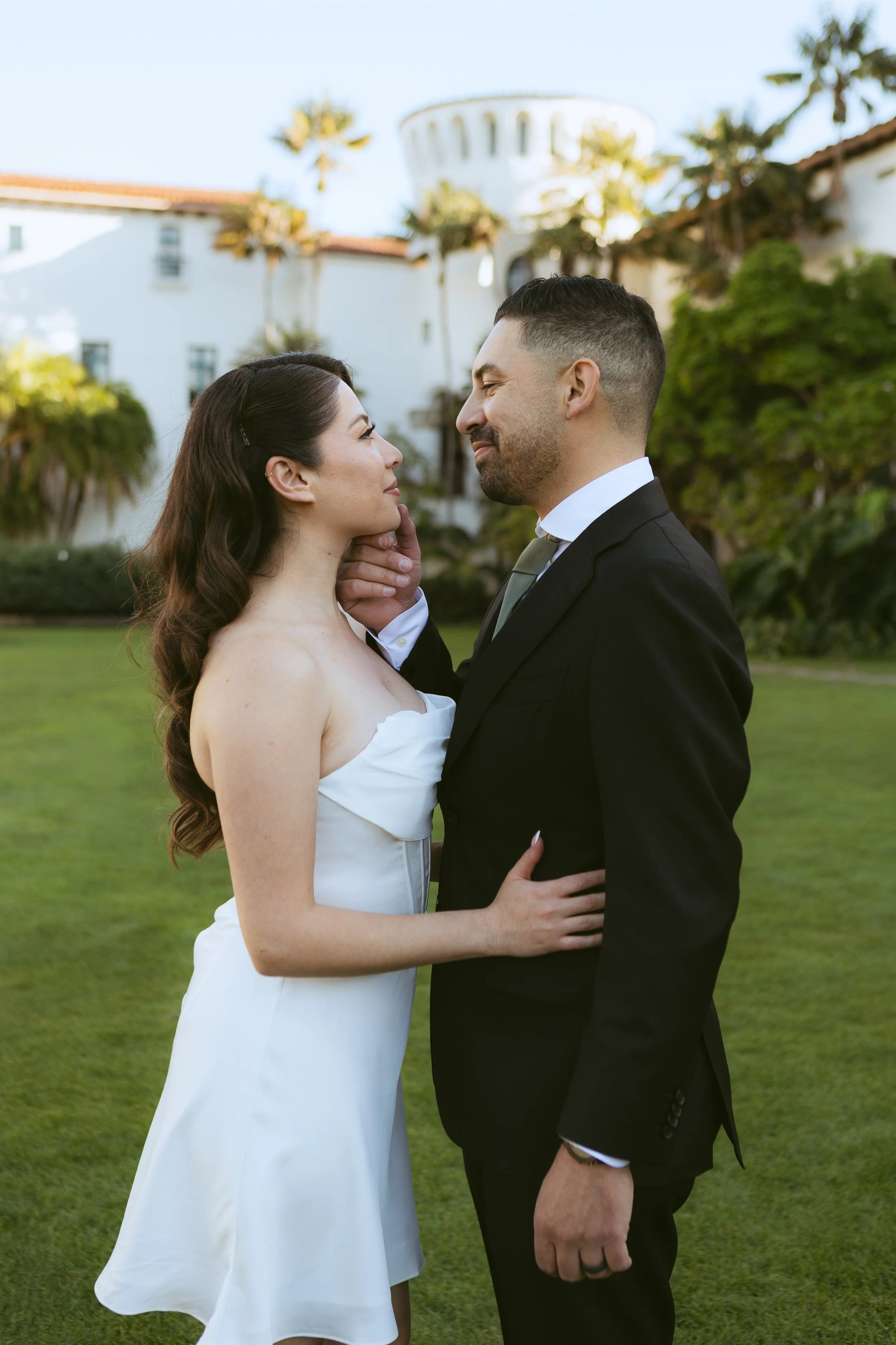 A couple in wedding attire looking into each other's eyes outdoors on a green lawn with palm trees and a white building in the background.