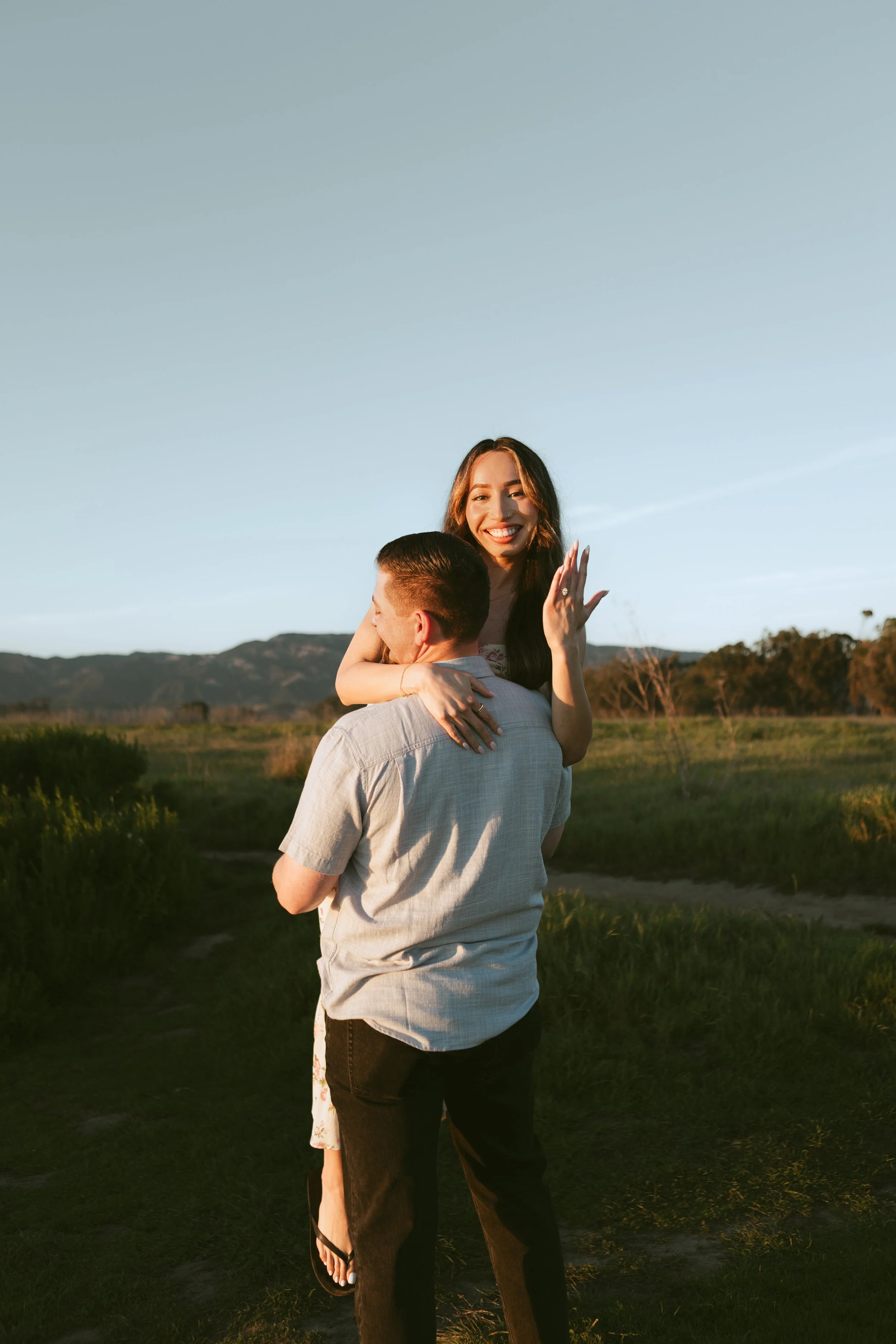A smiling woman with long dark hair being carried by a man in a field during sunset, with mountains in the background.
