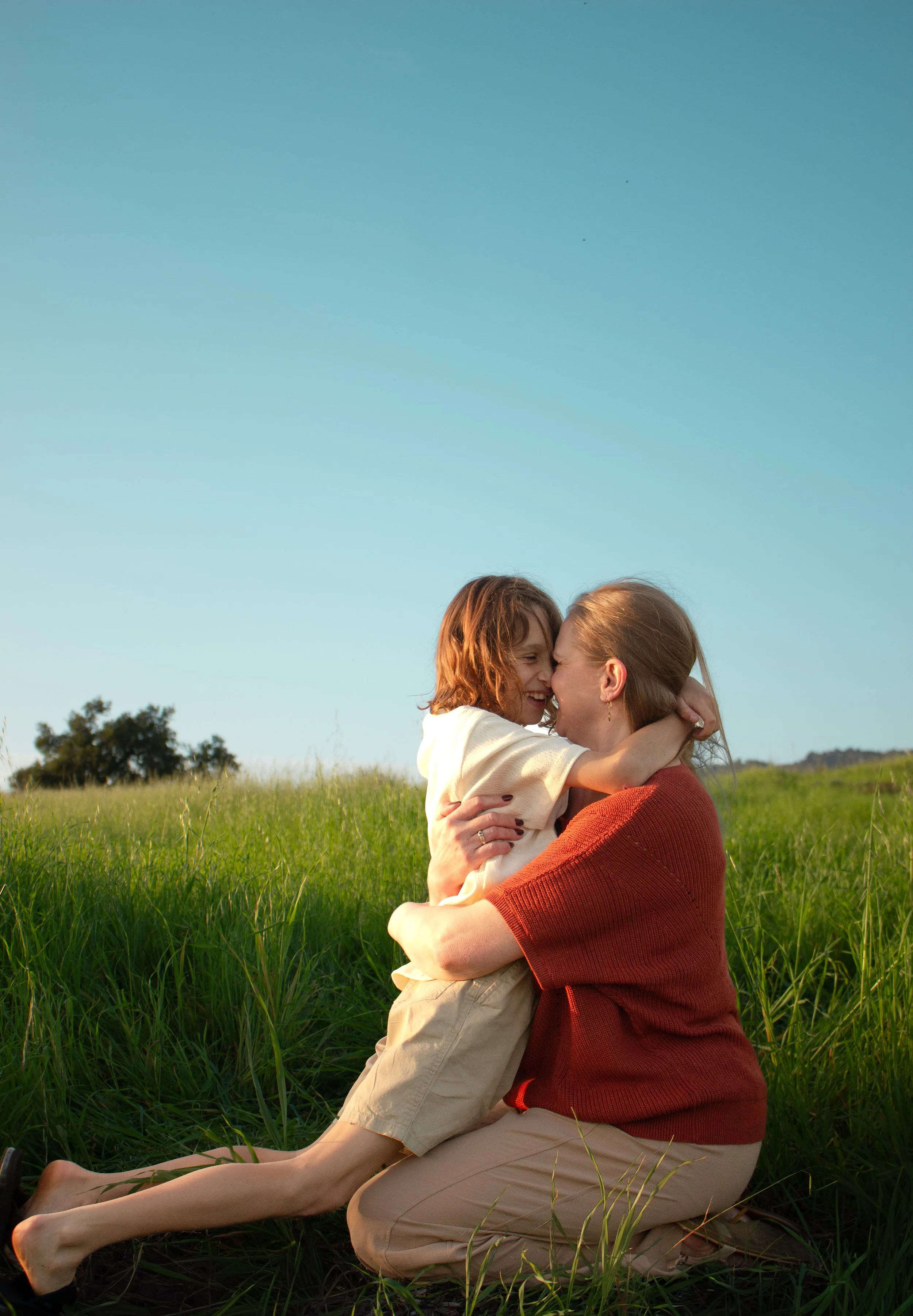 A woman and a child hugging in a grassy field under a clear blue sky in Santa Barbara, CA