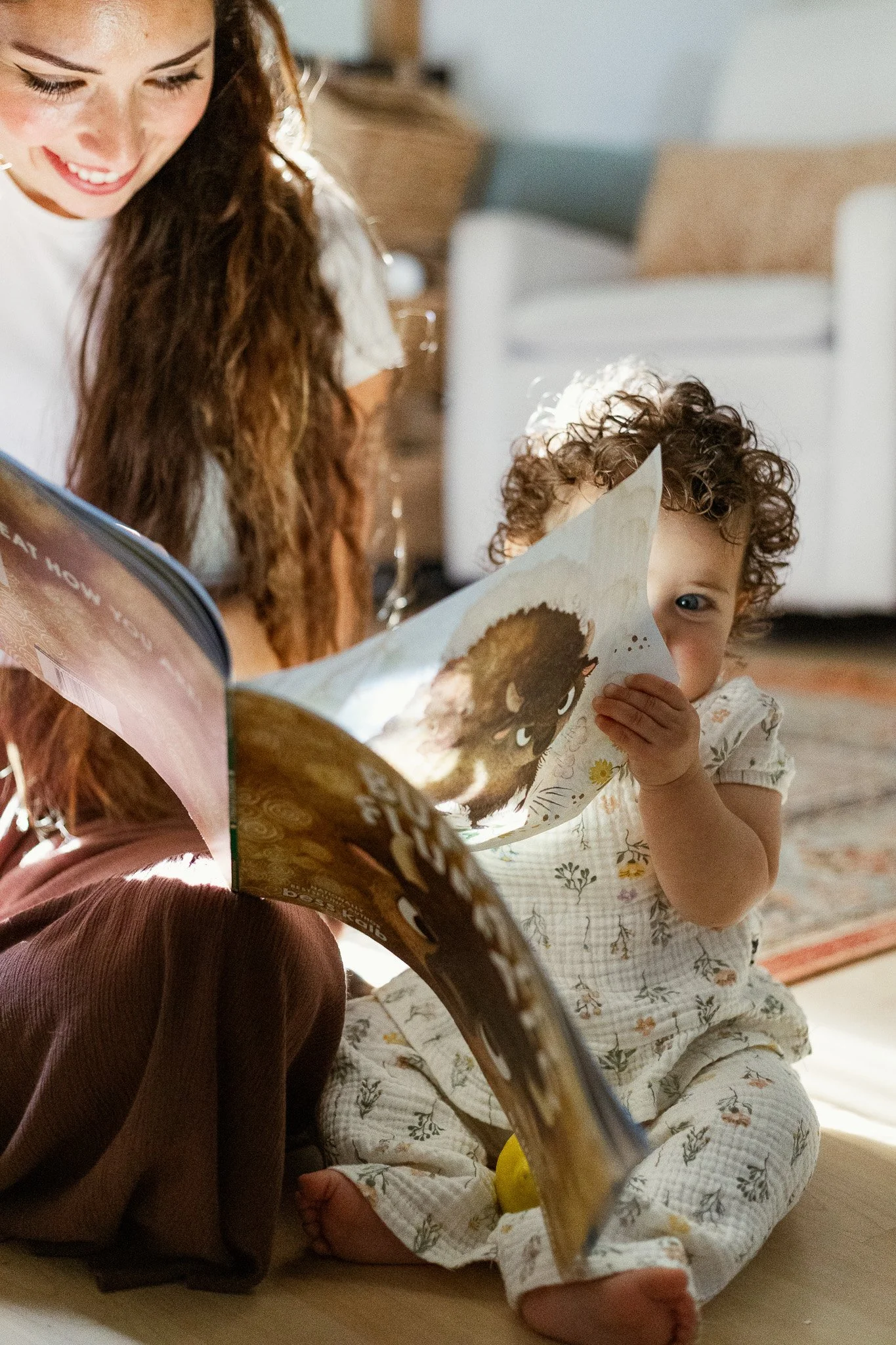 A woman and a young child sitting on the floor, reading a children's book together in a living room.