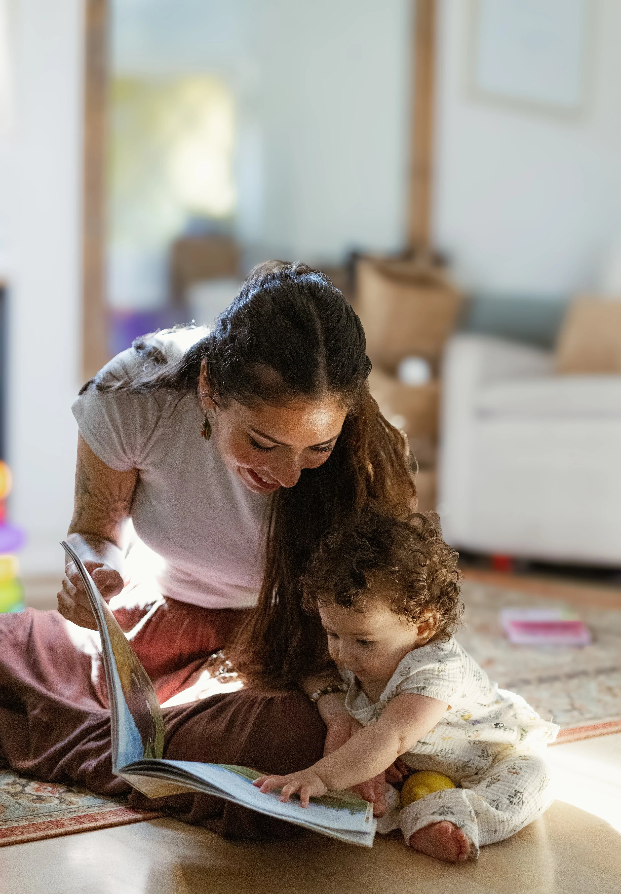 A woman and a young girl are lying on the floor together, looking at a book. The woman is smiling and the girl is reaching towards the book. The setting appears to be a cozy living room.
