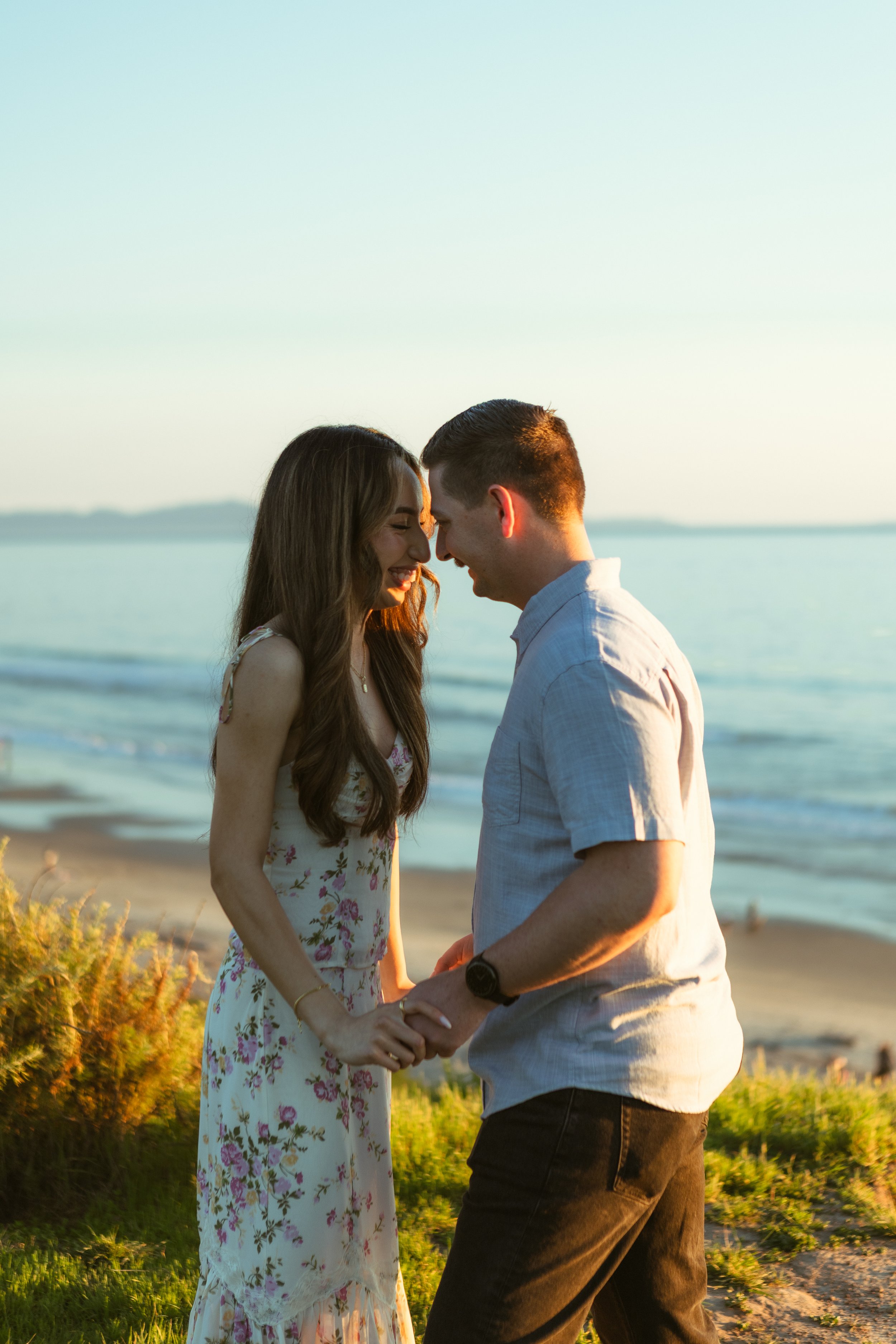 A couple holding hands and smiling at each other on the beach during sunset.