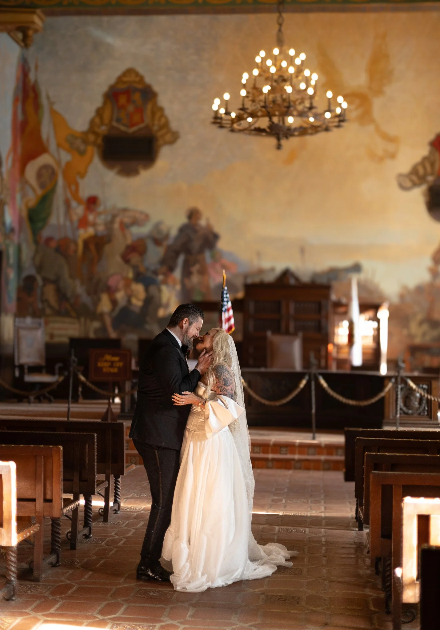 A bride and groom share a kiss inside a historic Santa Barbara courthouse decorated for a wedding, with a large chandelier, an American flag, and a mural on the wall.