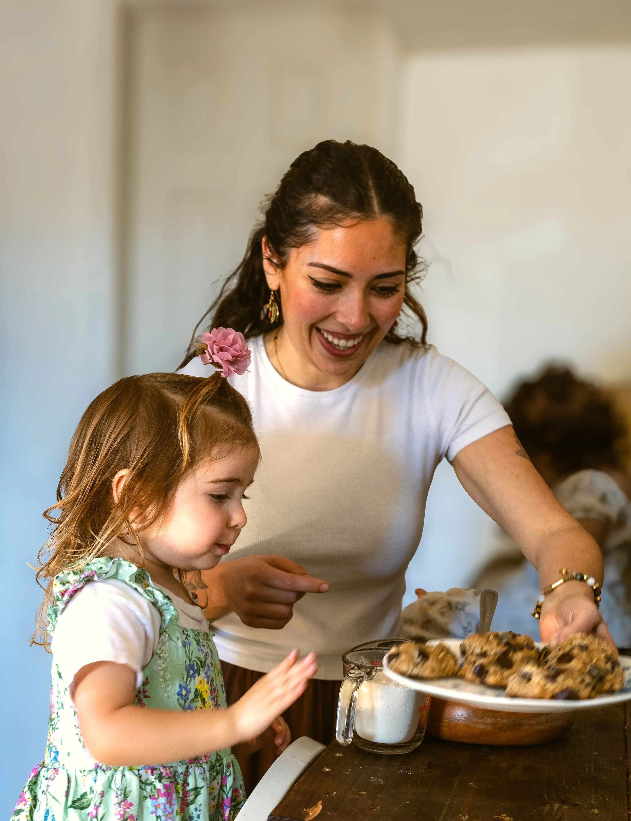 A smiling woman and a young girl are baking cookies together in a kitchen. The woman is reaching for cookies on a plate, while the girl watches closely at their home in Goleta, CA.