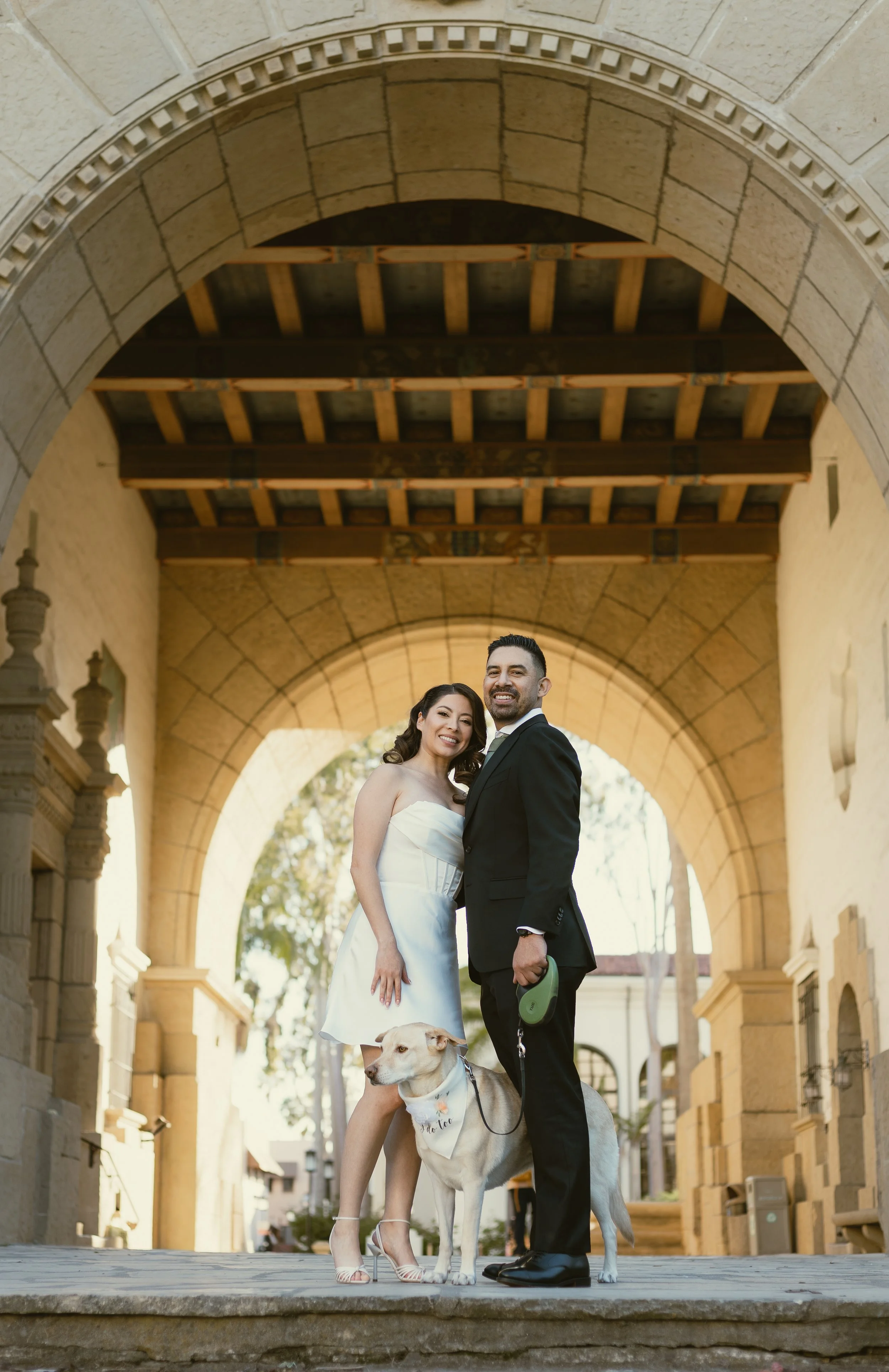 Smiling couple dressed in wedding attire, a woman in a white gown and a man in a black suit, standing under a large stone archway with a dog on a leash, in an outdoor historic setting.