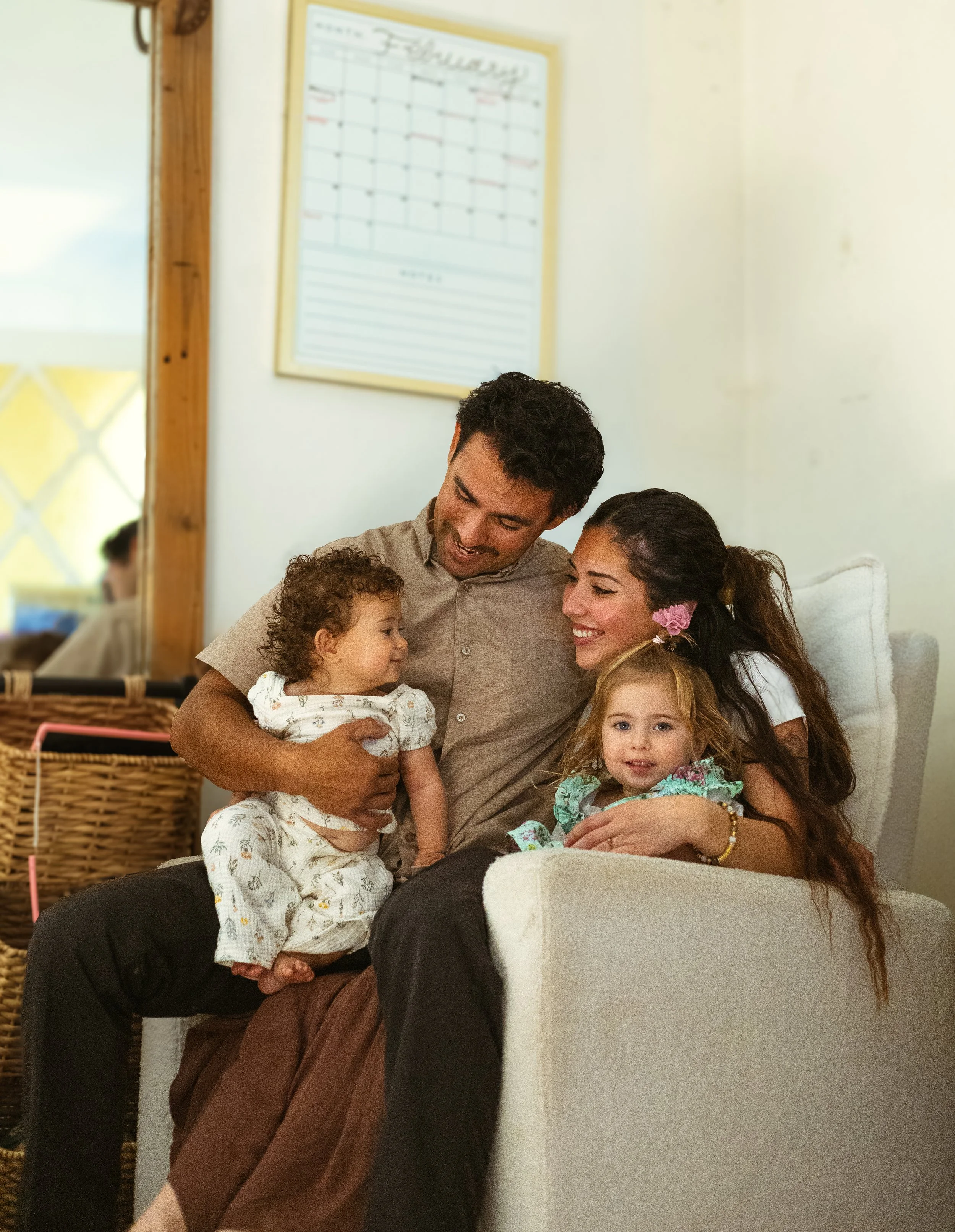 A family of four sitting together on a white couch, smiling, with two young girls and a man and woman, in a cozy living room in Goleta, CA