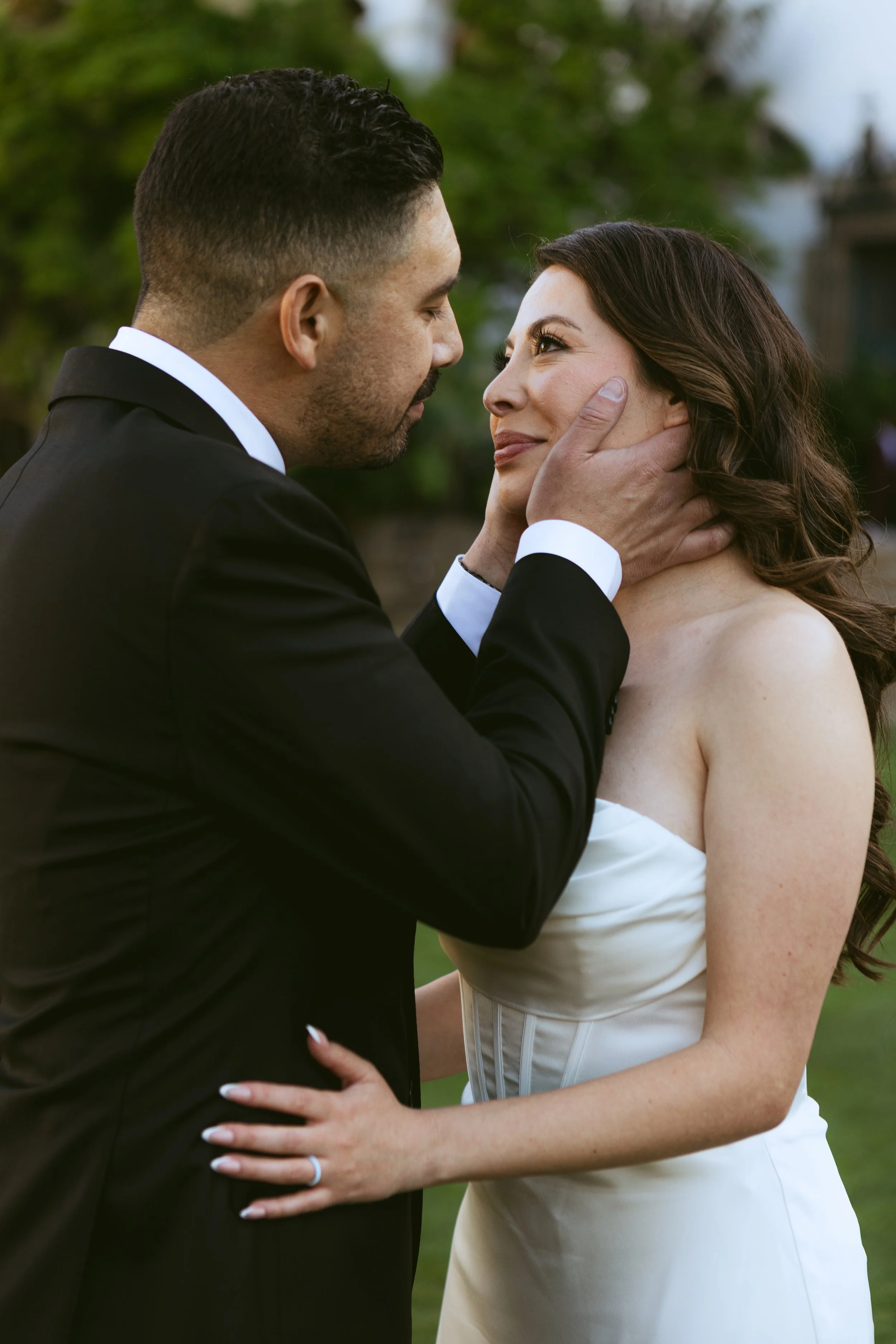A man in a black tuxedo gently holding a woman in a white wedding dress, about to kiss her during a wedding or romantic moment outdoors.