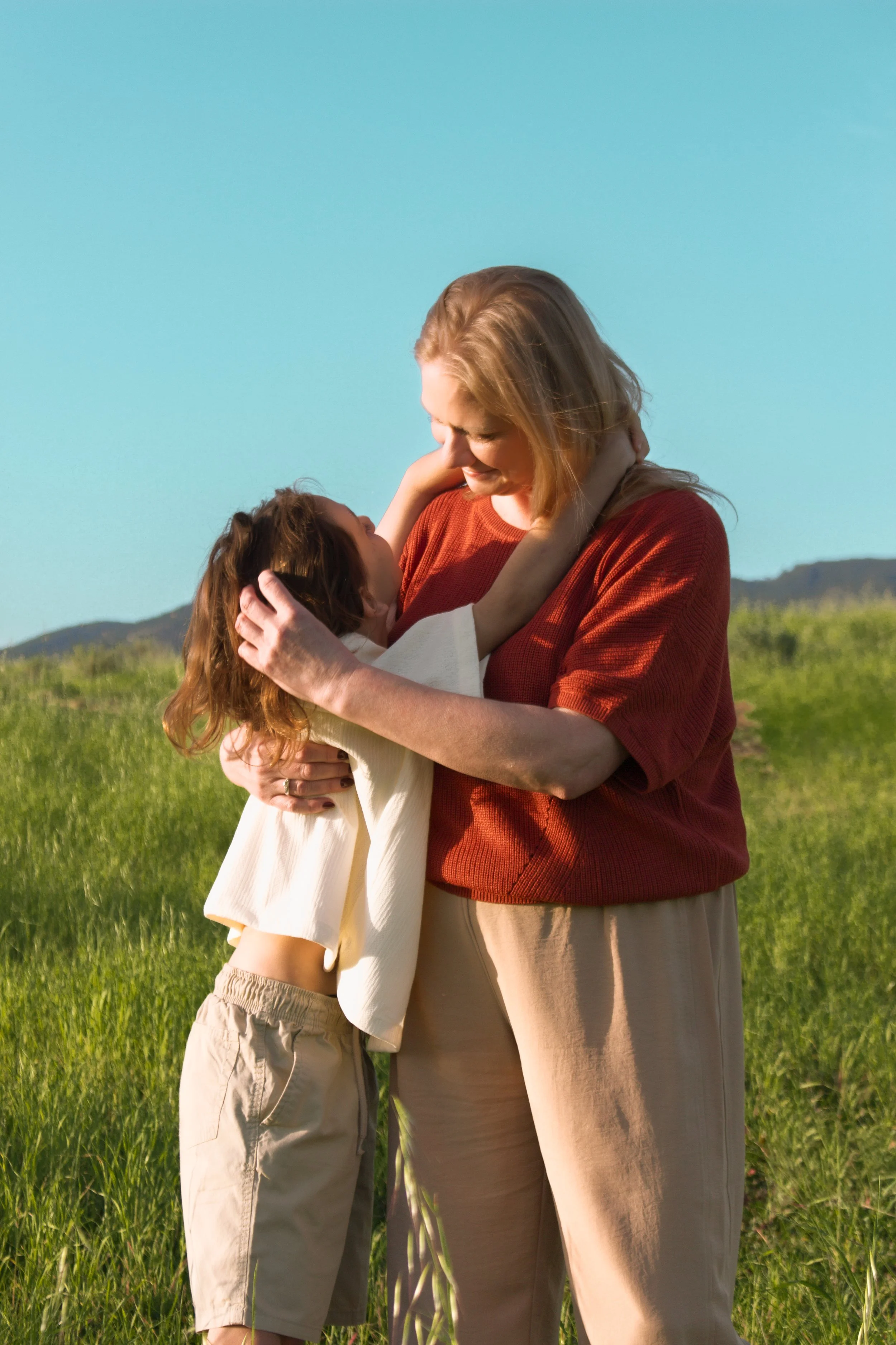 A woman and a young boy hugging outdoors on a sunny day in a field of green grass with mountains in the background.