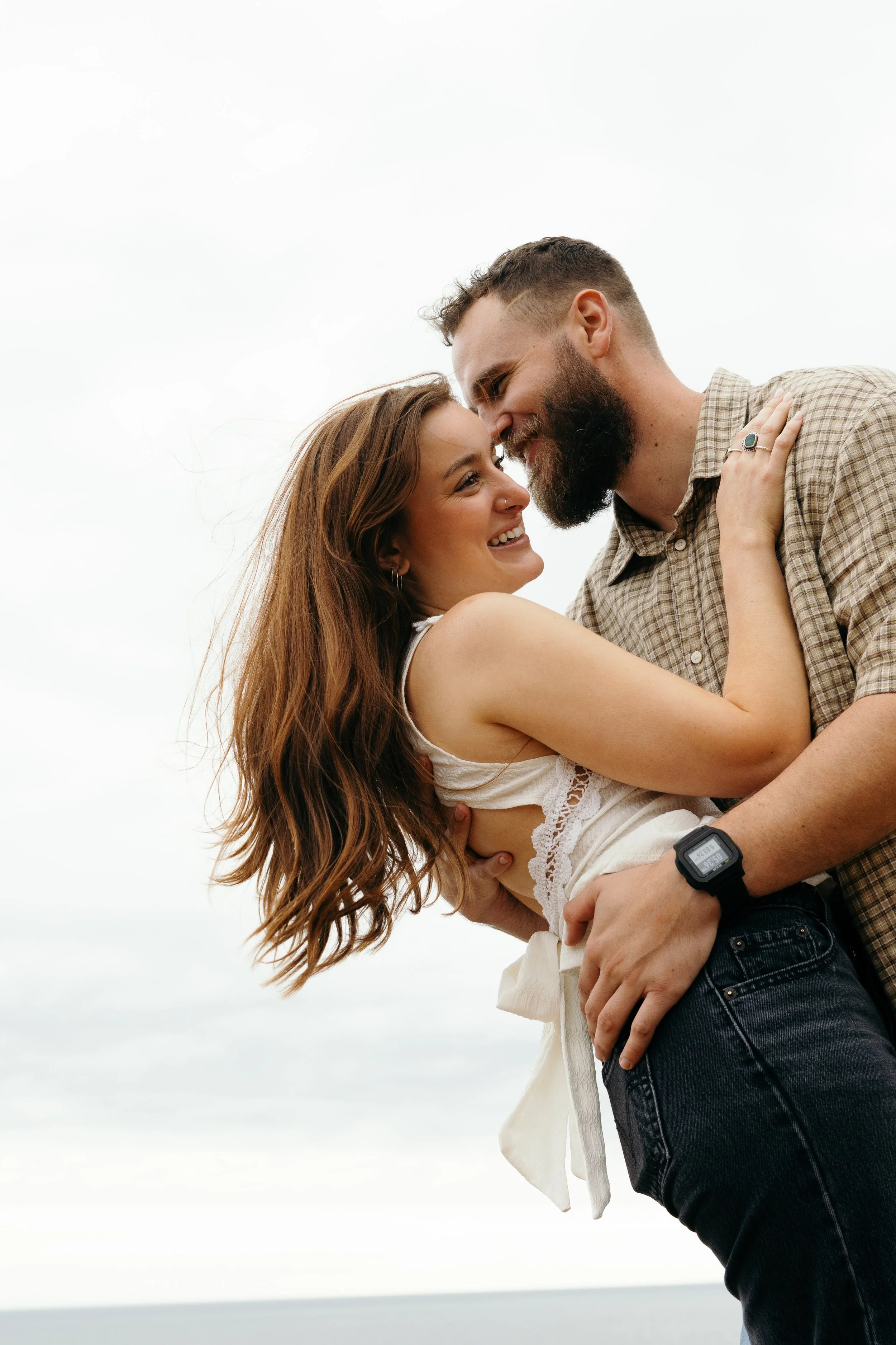 A man with a beard and a woman with long, wavy hair are embracing outdoors against a cloudy sky. They are smiling and touching foreheads with their eyes closed, appearing happy and affectionate.