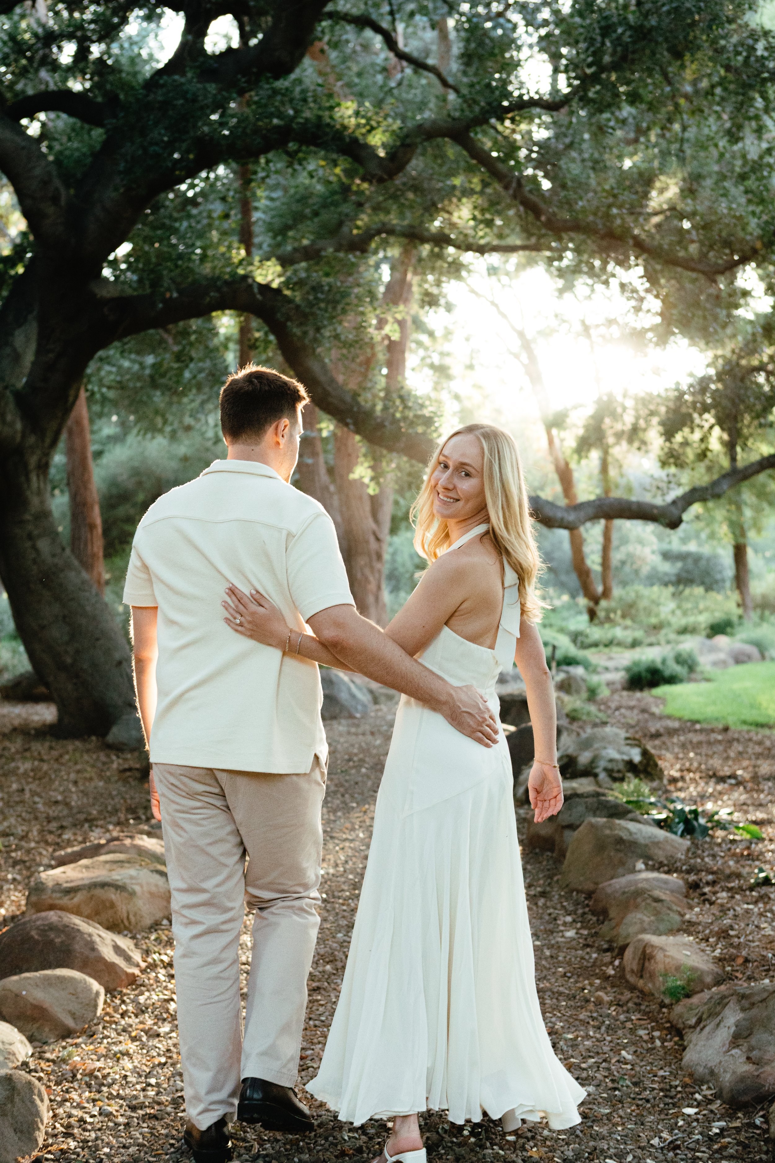 A couple stands outdoors in a forested area with large trees and rocks, holding hands and looking at each other, during sunset.