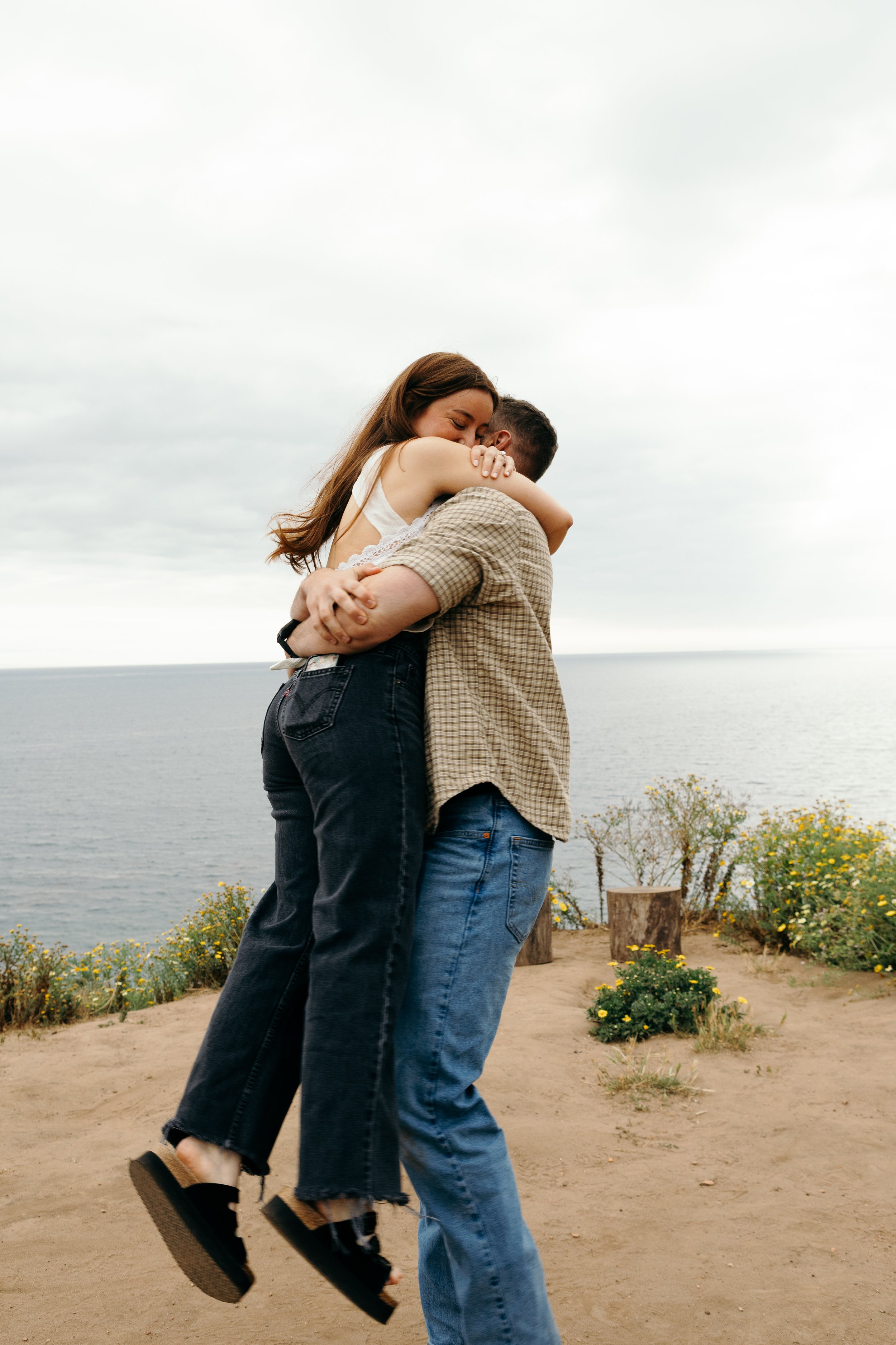 A woman with long brown hair and a man with short dark hair embracing outdoors near the water, with yellow flowers and wooden logs in the background.