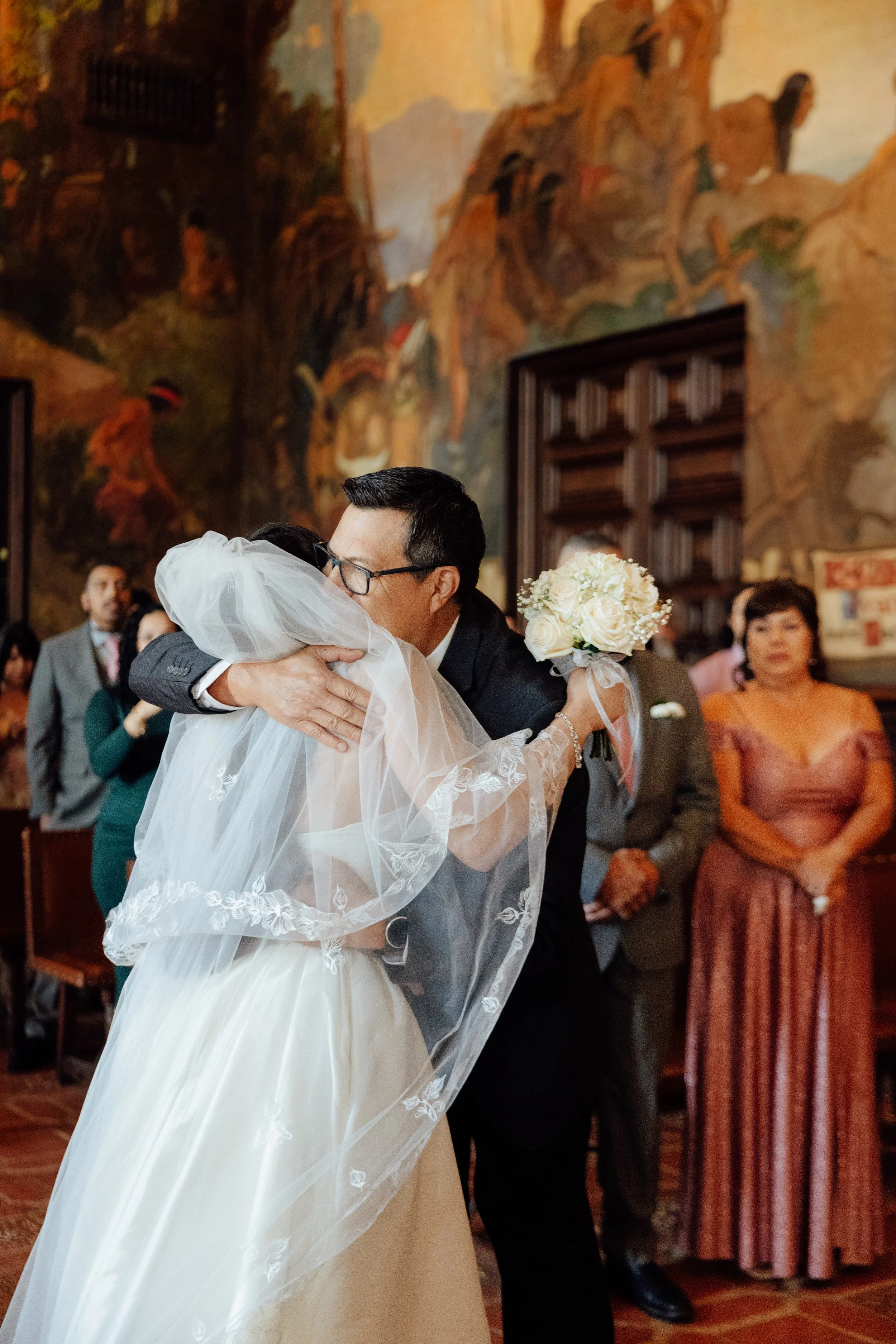 A bride and a groom hugging during a wedding ceremony inside a decorated church. The bride wears a white wedding gown with lace details and a veil, and holds a bouquet of white roses. The groom wears a dark suit and glasses. Guests are visible in the background.