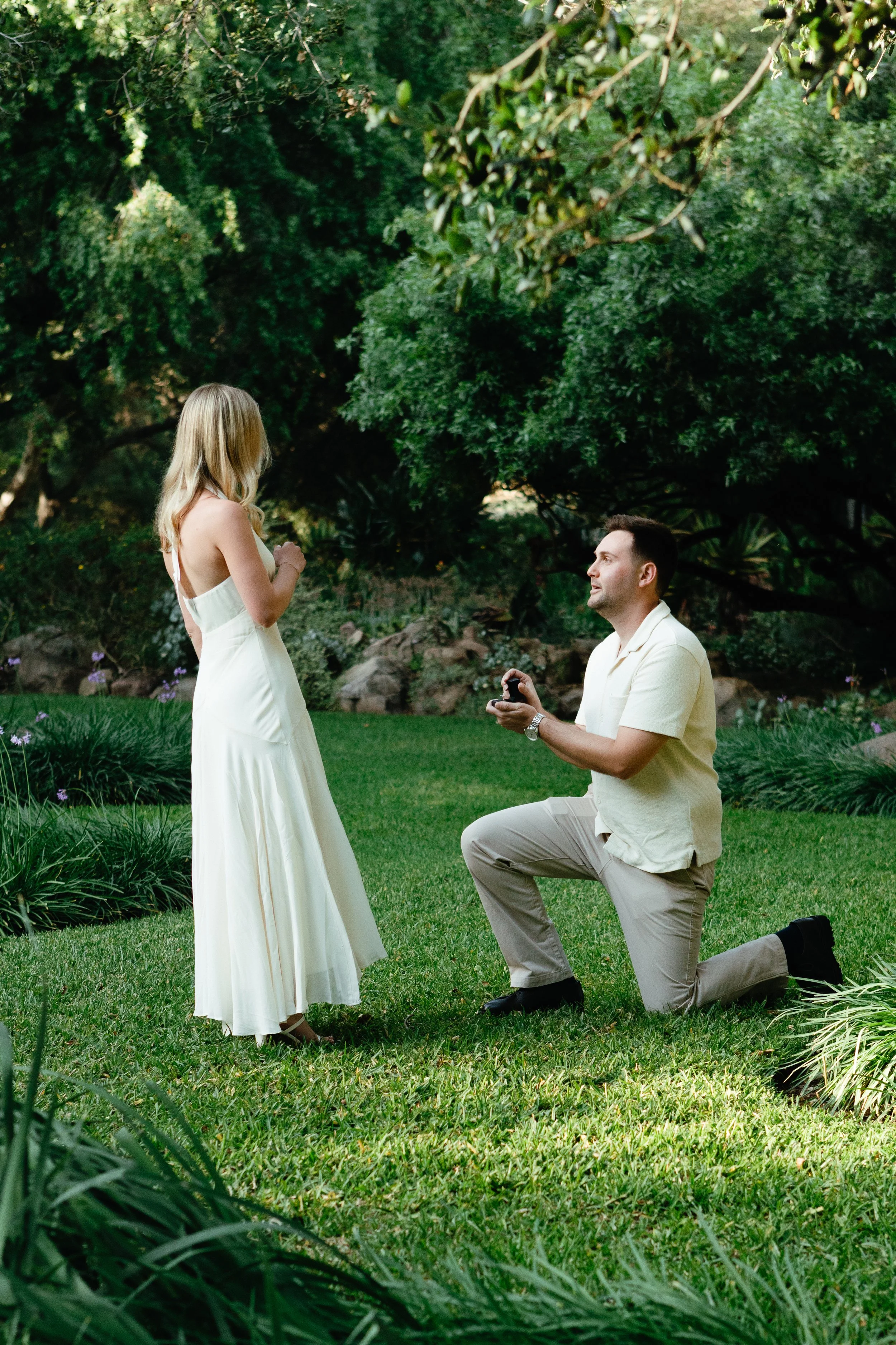 A man kneeling on one knee proposing to a woman in a garden with green trees and grass, during daytime.