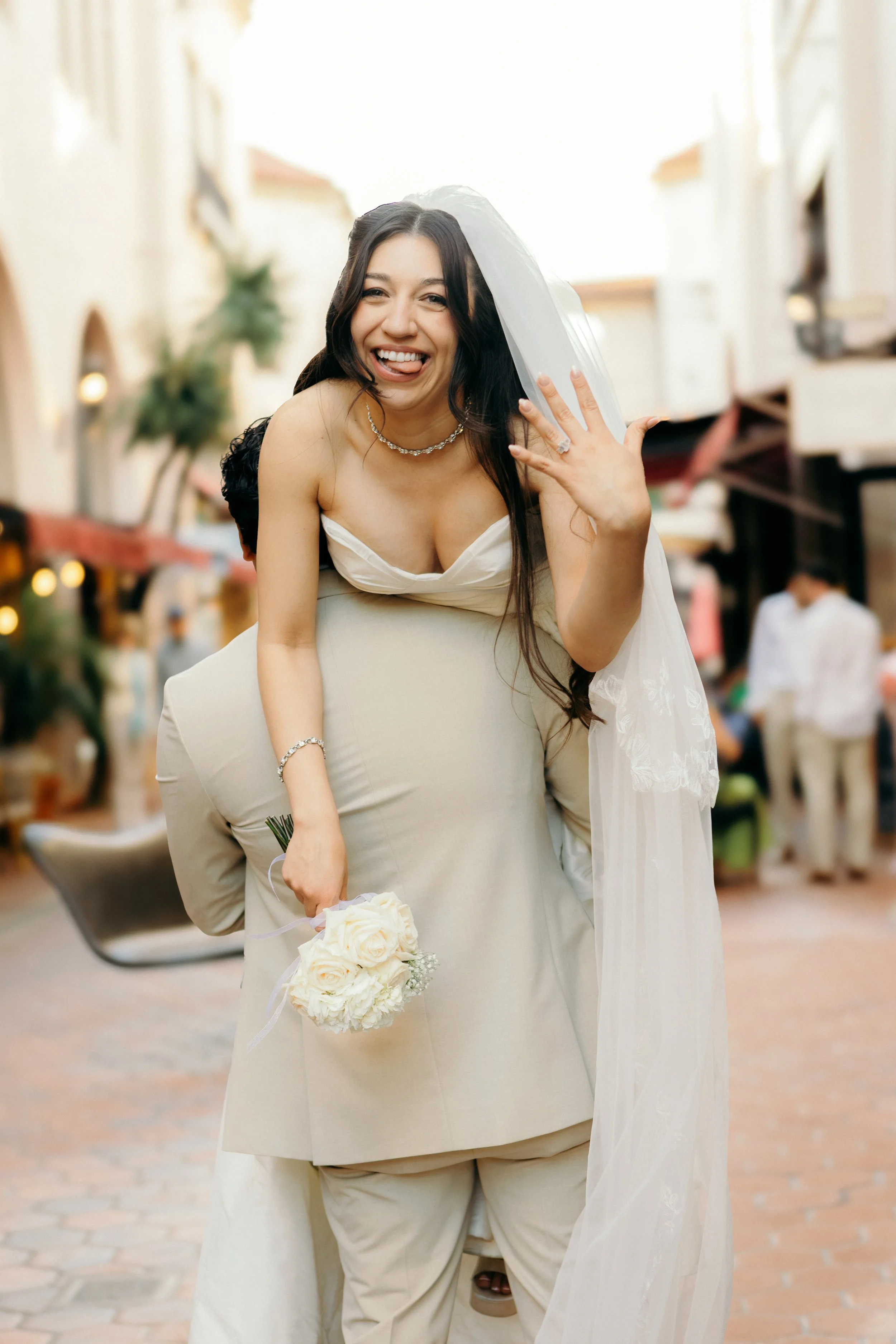 A bride waving and smiling, with her tongue sticking out, holding a bouquet of white roses. She is wearing a white wedding dress, a veil, and jewelry. People are blurred in the background of an indoor, warmly lit venue.
