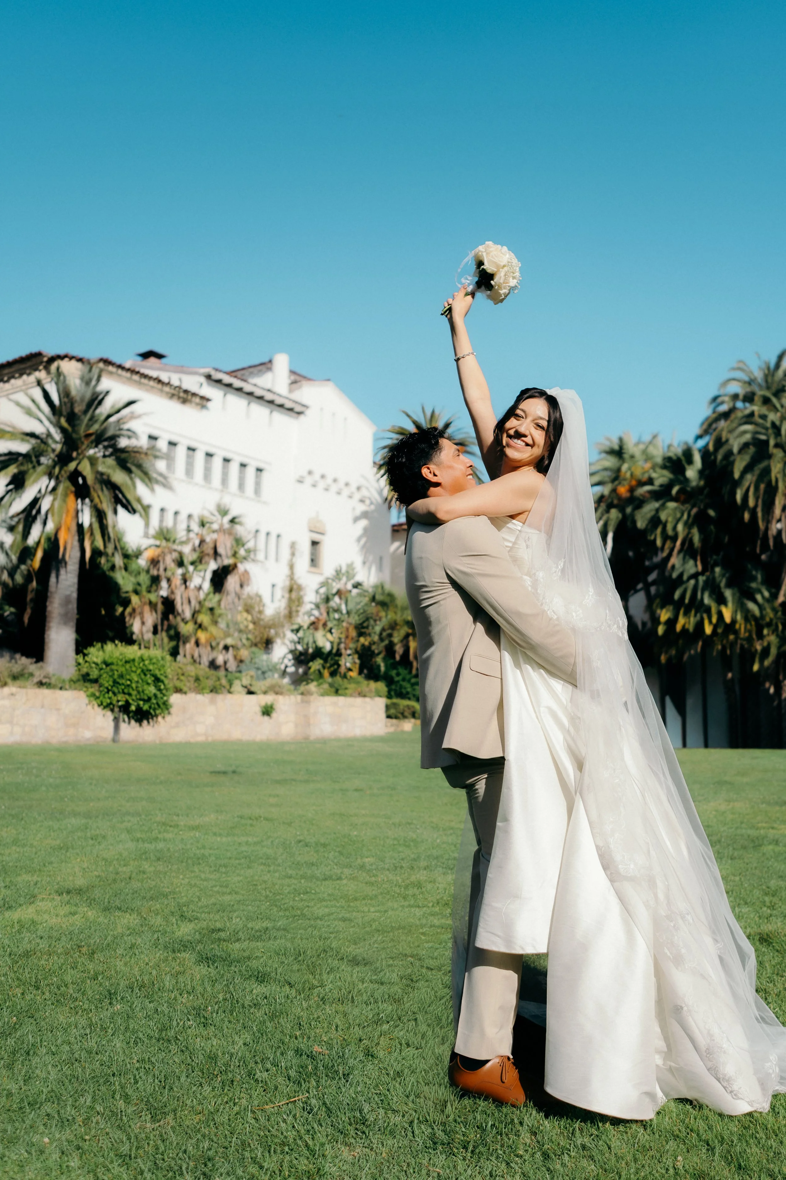A couple dressed in wedding attire, with the groom lifting the bride in his arms on a grassy lawn, holding a bouquet, with palm trees and a large white building in the background under a clear blue sky.