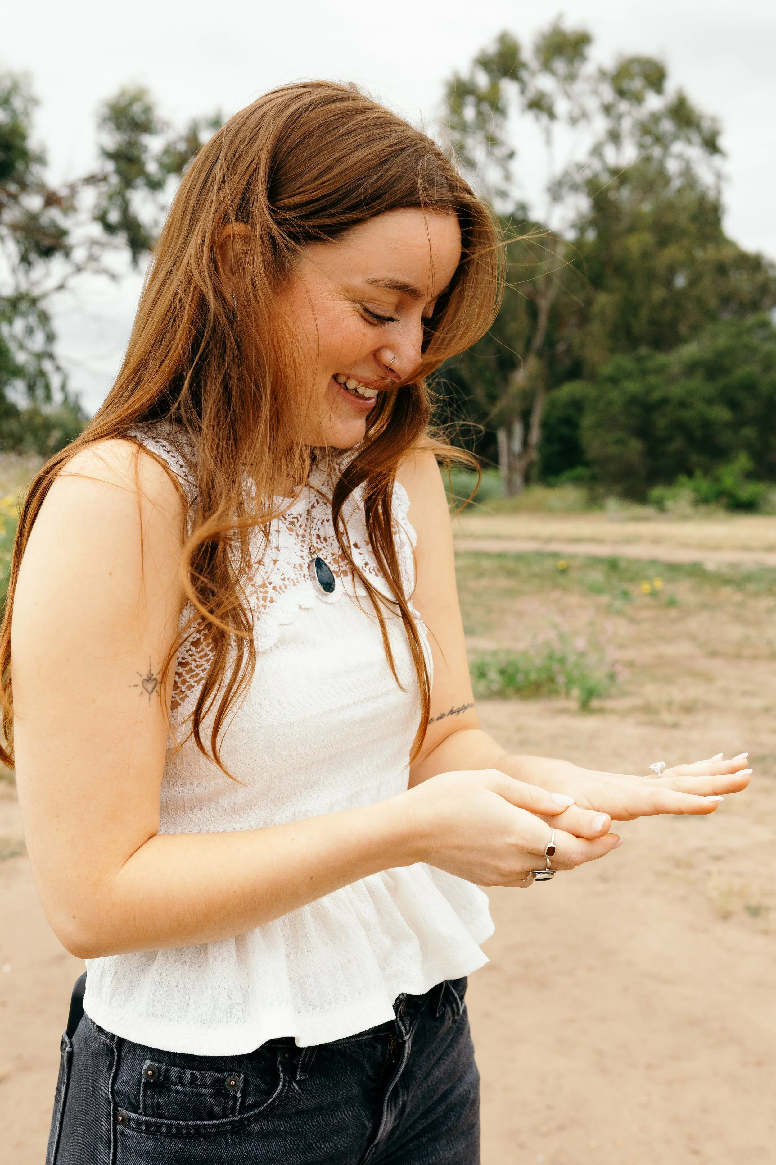 A young woman with long, wavy red hair and a nose piercing is smiling while looking down at her hands, which are cupped together. She is wearing a white sleeveless top with lace detail and a black pendant necklace, outdoors with trees and a cloudy sky in the background.