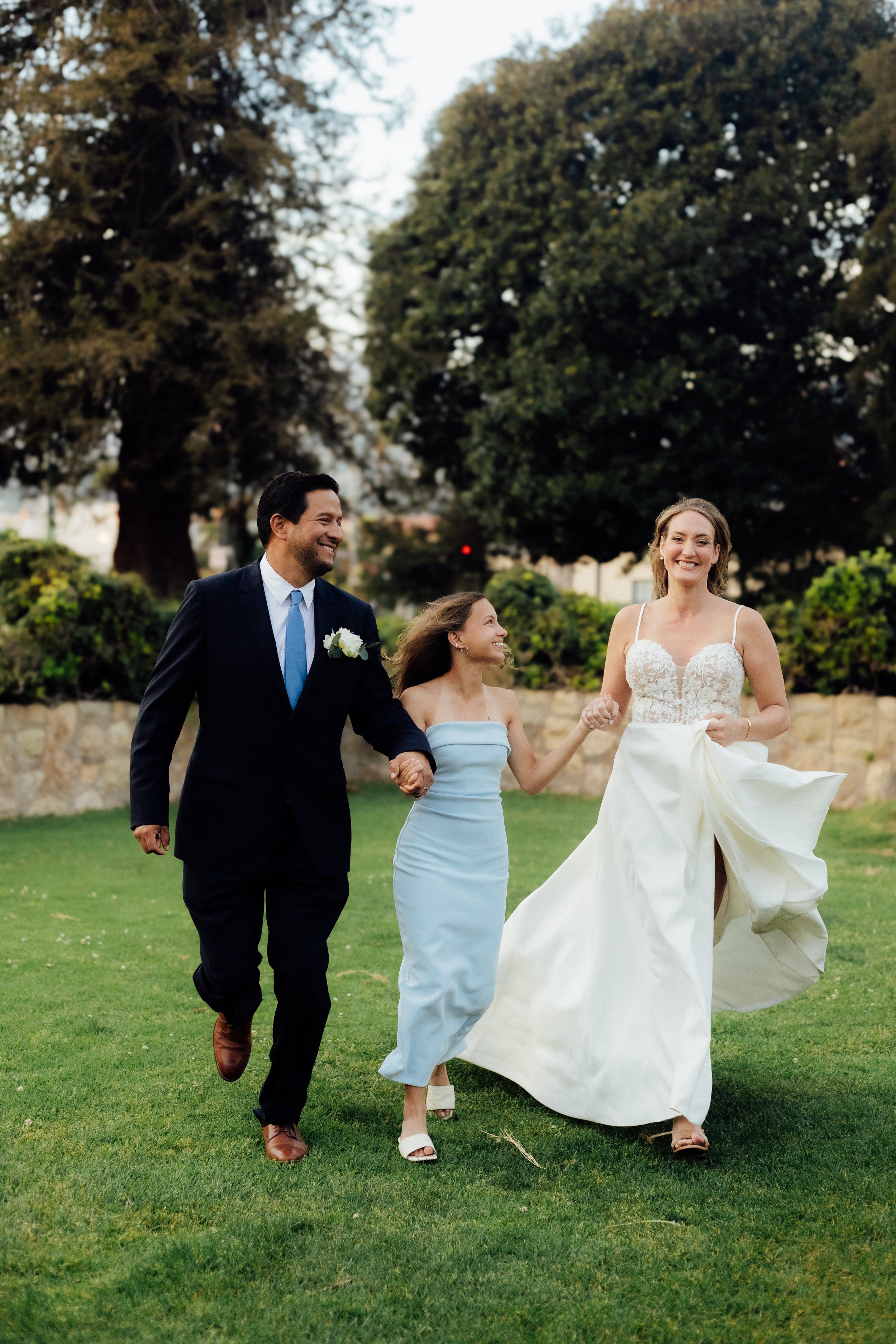A groom, a bride, and a bridesmaid walking outdoors on a green lawn with large trees in the background. The groom is wearing a navy suit, white shirt, and a blue tie, holding hands with the bride. The bride is wearing a wedding gown, smiling, holding her dress with one hand. The bridesmaid is in a light blue dress, smiling, holding the bride's hand.