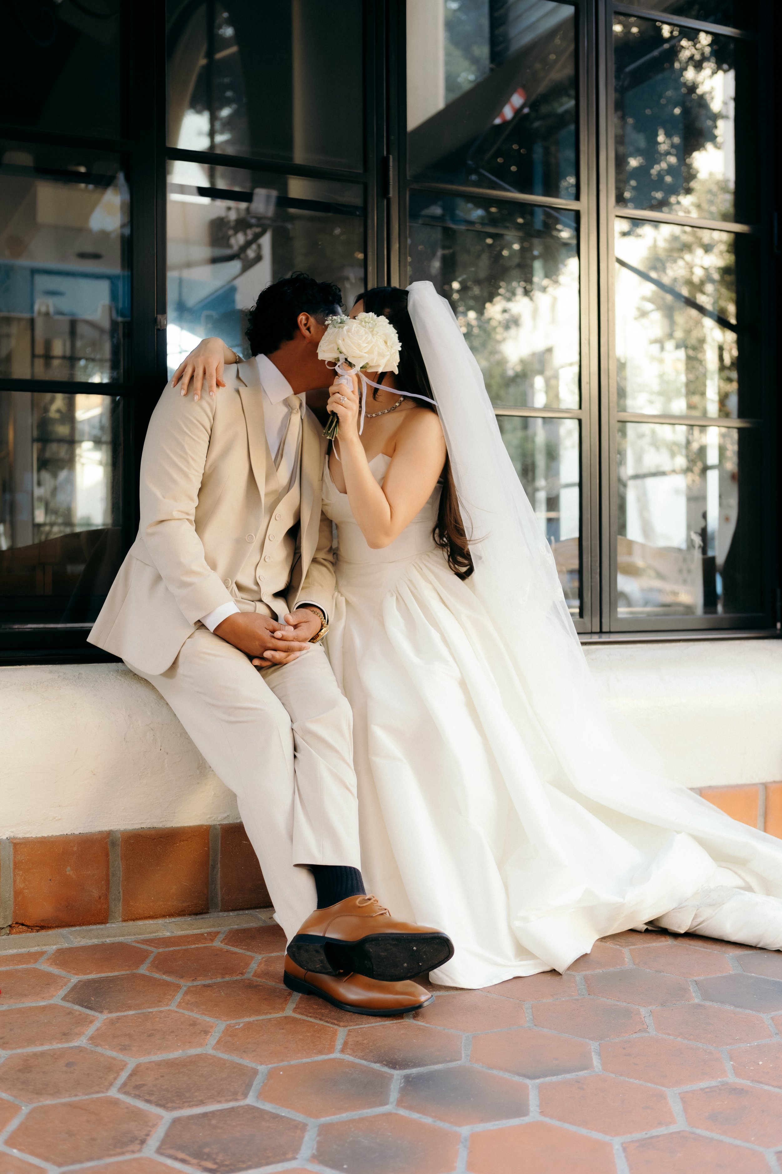 A bride and groom are sitting on a ledge outside near a glass building. The bride, wearing a white wedding gown and veil, is holding a bouquet of white flowers and partially covering her face. The groom, in a cream-colored suit and brown shoes, is le