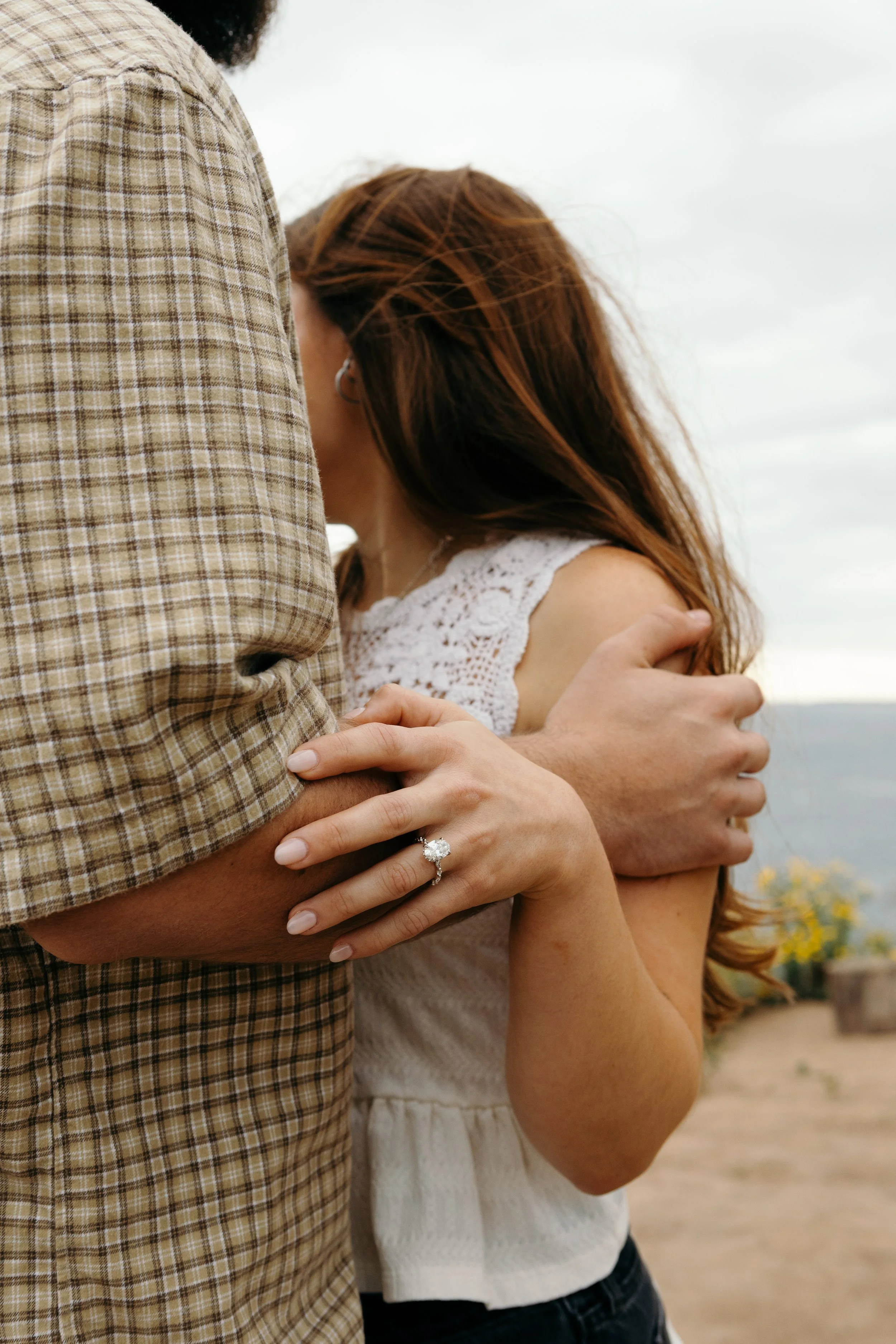 A man and woman embracing outdoors, with the man wearing a plaid shirt and the woman wearing a white lace top, showing a diamond ring on her finger.