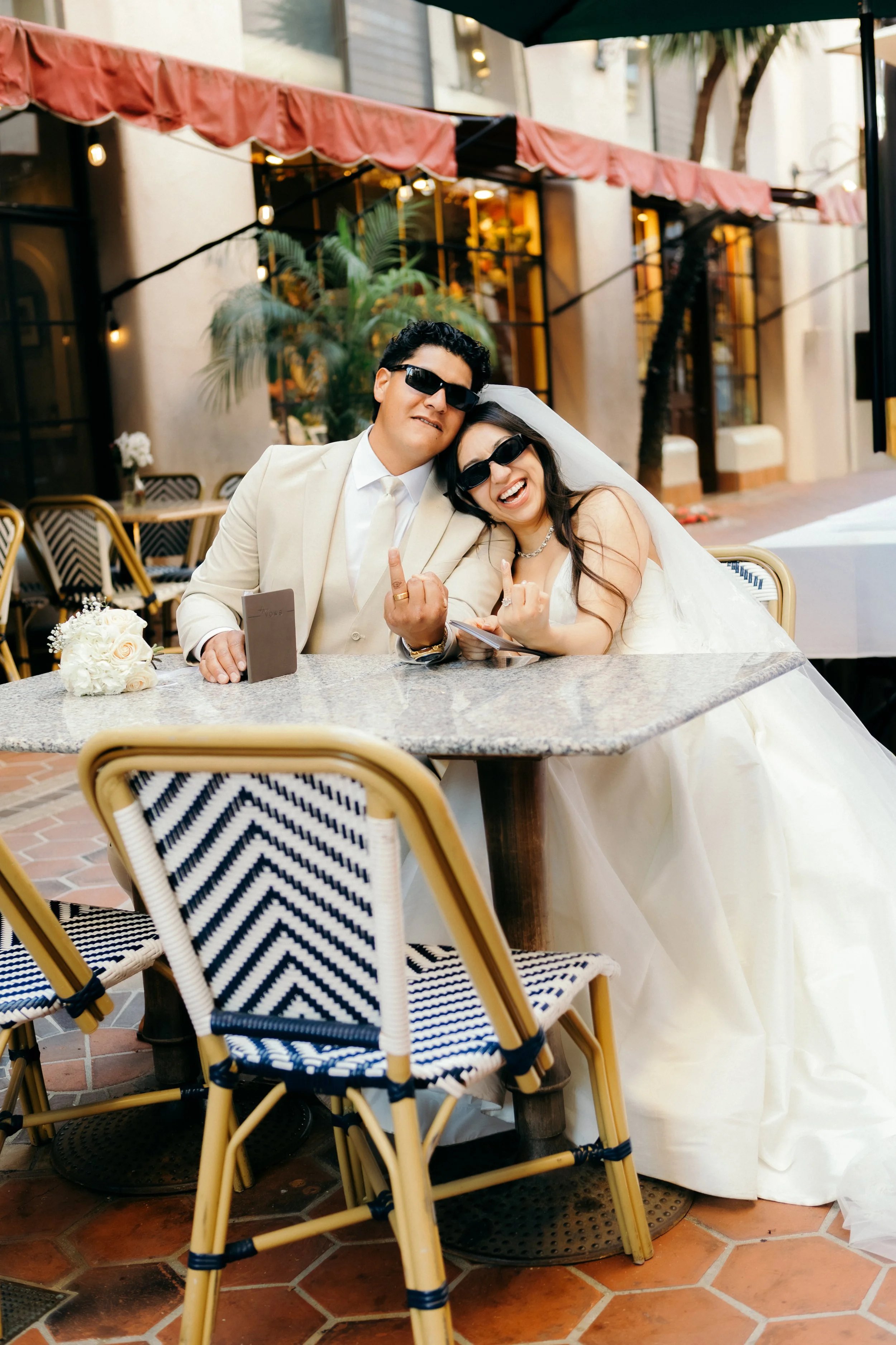 A newlywed couple in formal wedding attire, wearing sunglasses, sitting at a table in an outdoor restaurant or cafe with a pink awning and decorative chairs.