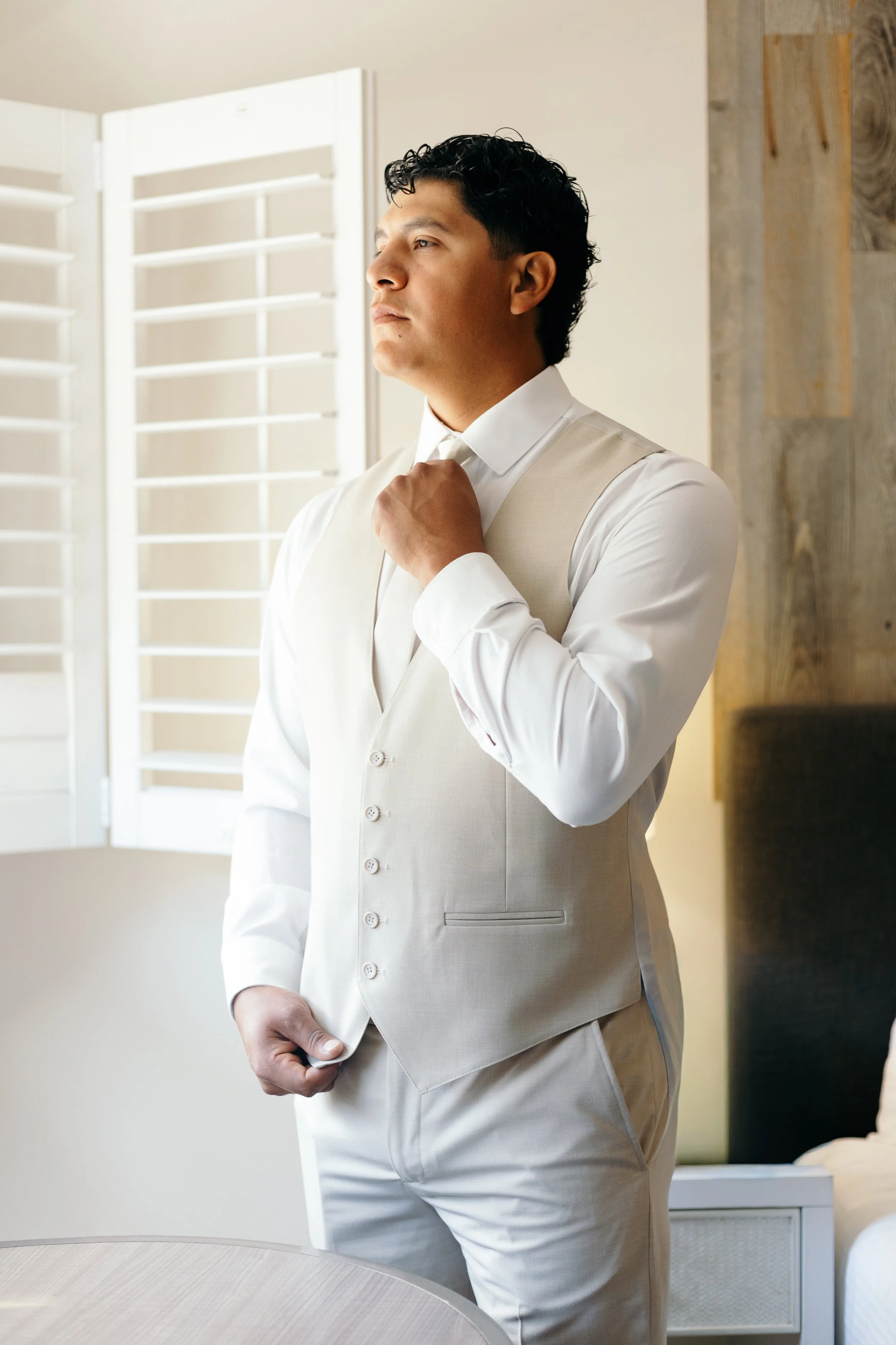 A man in a beige suit adjusting his tie in a room with a window, a round table, and a bed.