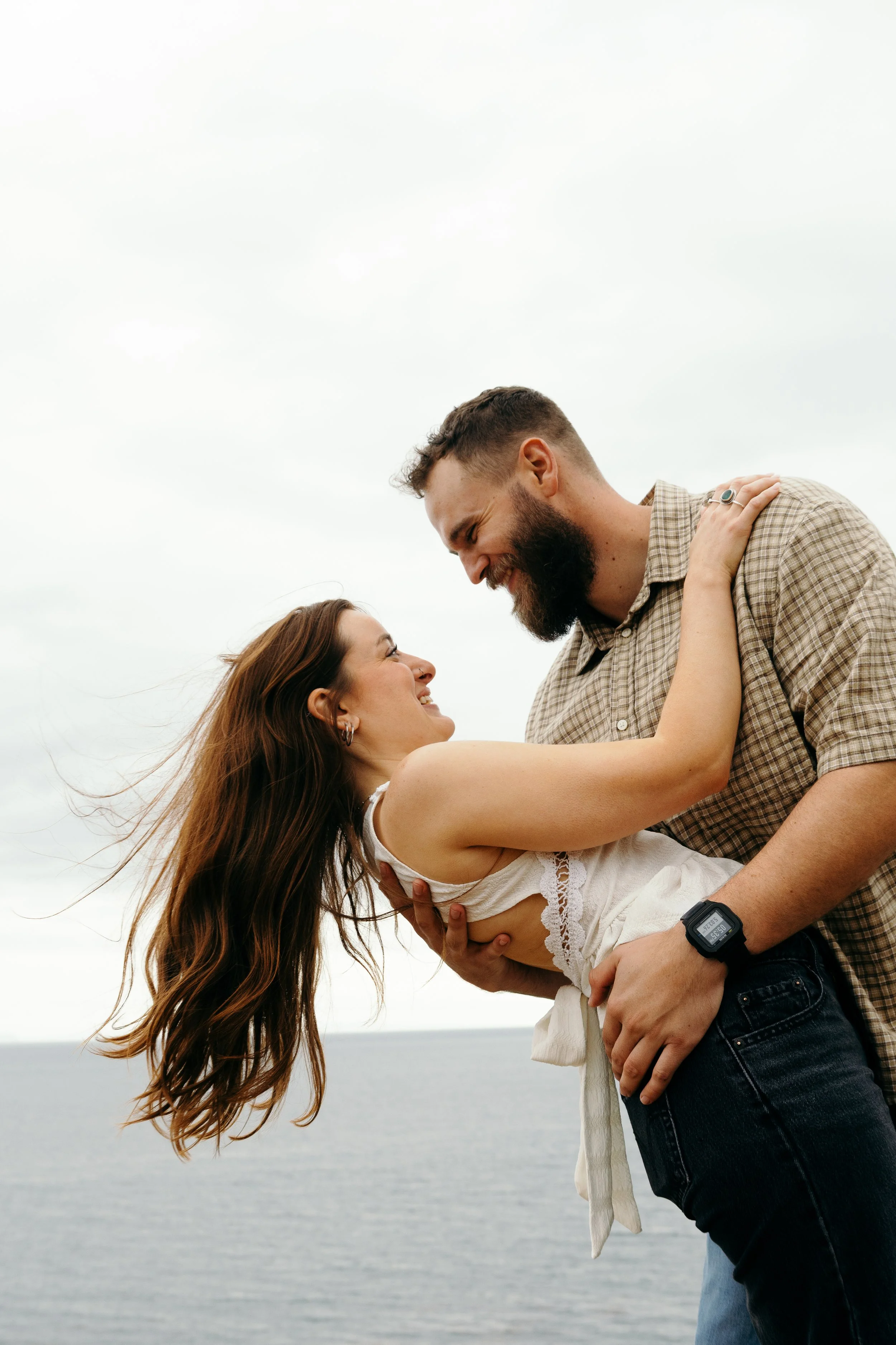 A couple is outdoors near a large body of water, with the woman being lifted by the man. They are smiling and looking at each other with a cloudy sky overhead.