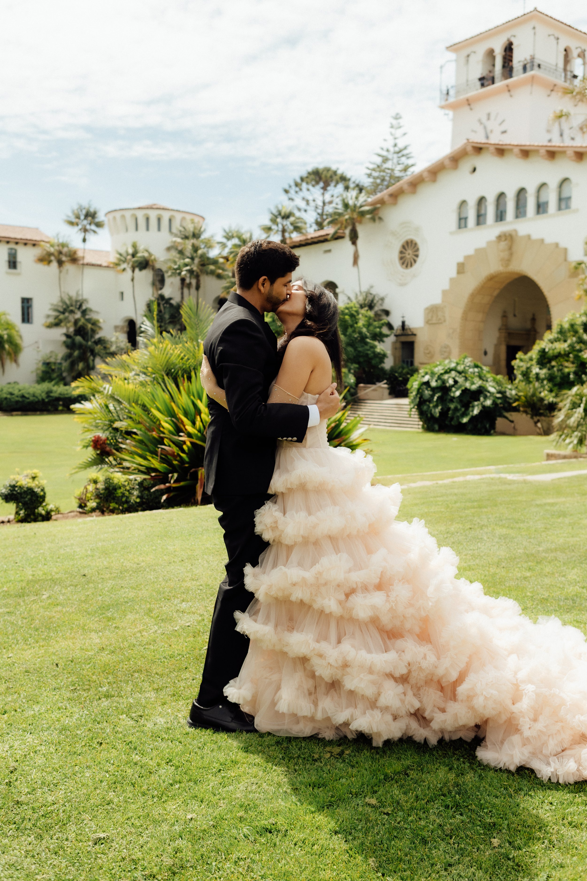 A couple kissing outdoors on a wedding day, with a grand white building and palm trees in the background.