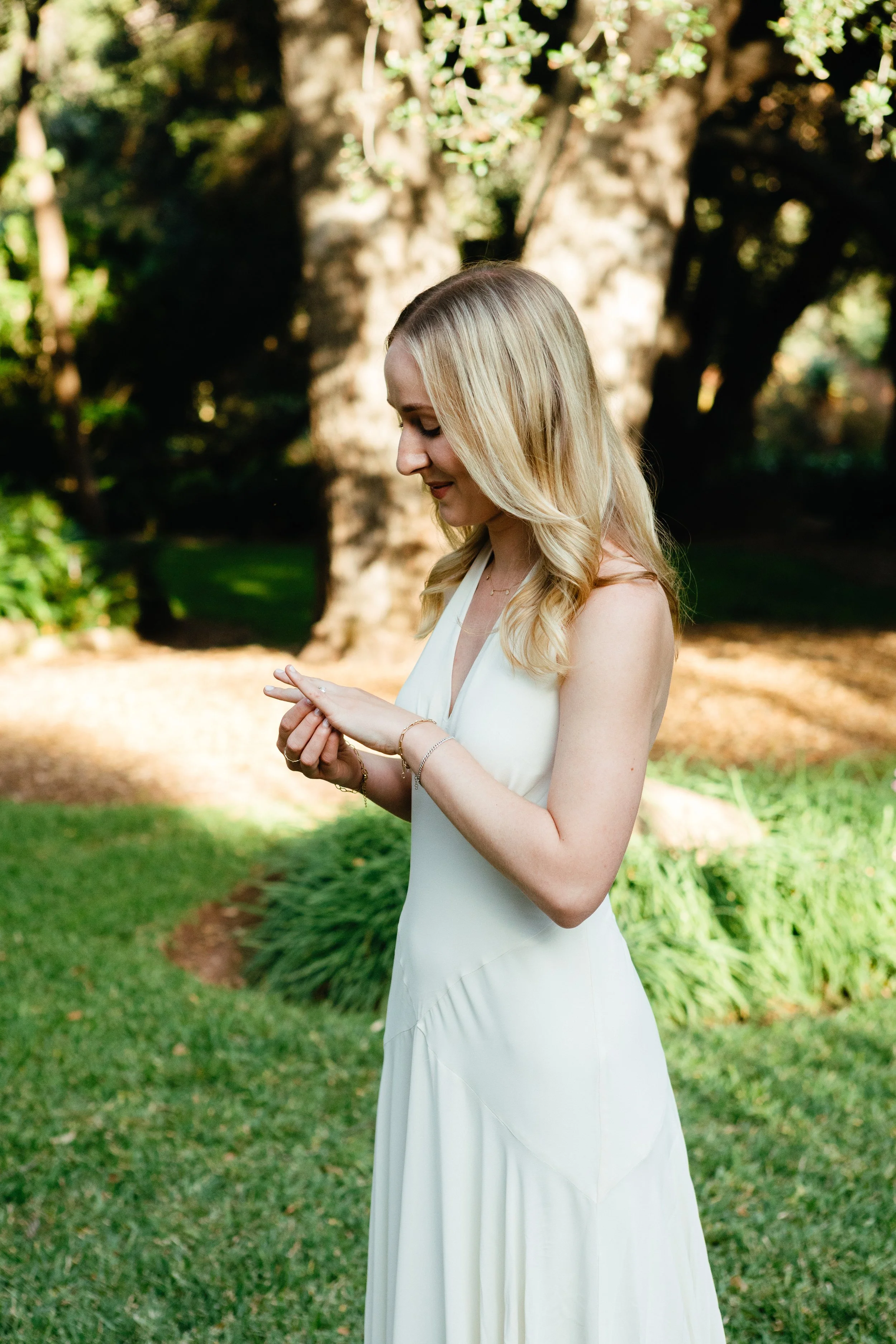 A woman with long blonde hair wearing a white dress standing outdoors in a garden. She is looking down and clasping her hands, with a large tree and lush greenery in the background.