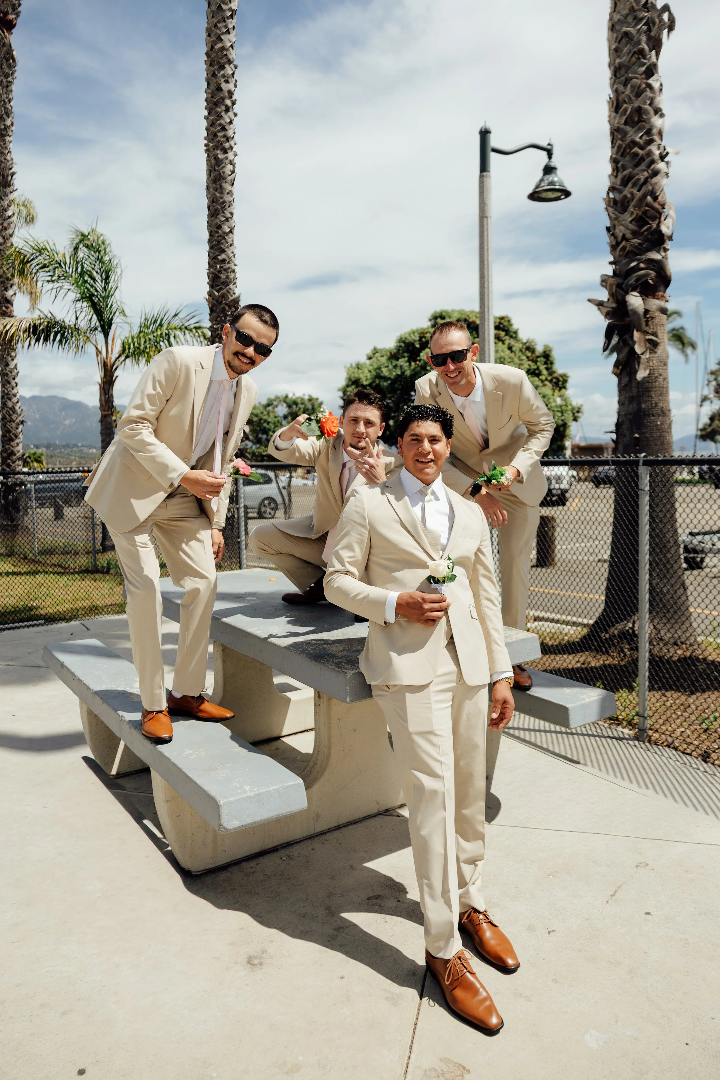 Five men dressed in beige suits and brown shoes, some wearing sunglasses, posing playfully outdoors near palm trees under a partly cloudy sky.