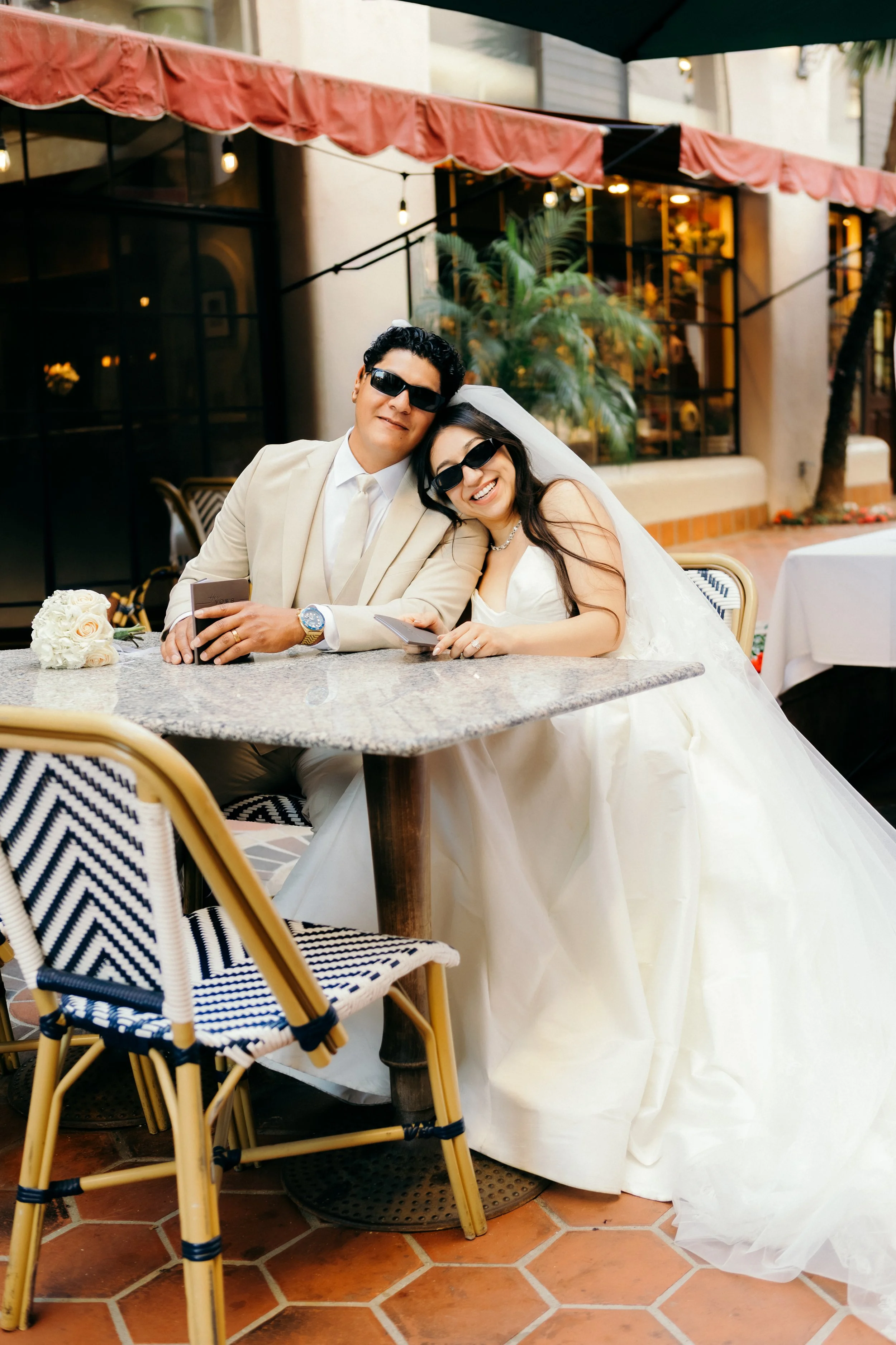A bride and groom sitting at a table outdoors, both wearing sunglasses and smiling. The bride is in a white wedding dress with a veil, and the groom is in a cream suit with a white shirt and tie. They are holding hands and leaning towards each other. The table has a bouquet of white roses, and there are chairs with black and white striped cushions. The background shows a building with large windows, some greenery, and string lights.