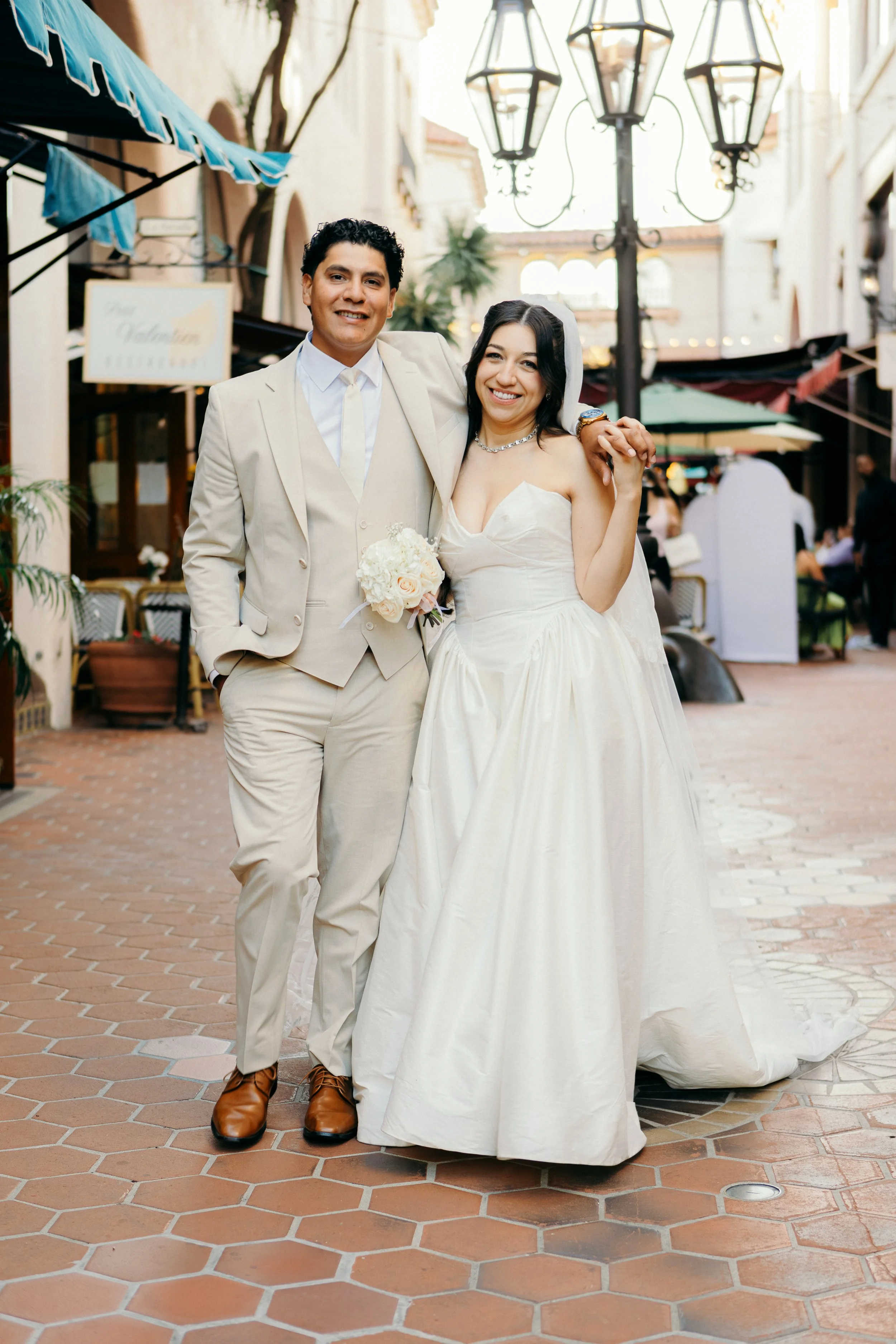 A smiling bride and groom are standing close together outdoors in a European-style street setting. The groom is dressed in a light beige suit with a white shirt and beige tie, holding a bouquet of white roses, and has dark curly hair. The bride is wearing a strapless white wedding gown with a sweetheart neckline, a pearl necklace, and a veil, with dark wavy hair. They are holding hands, and the groom's arm is around the bride's shoulders. There are decorative street lamps and buildings with arches and balconies in the background.