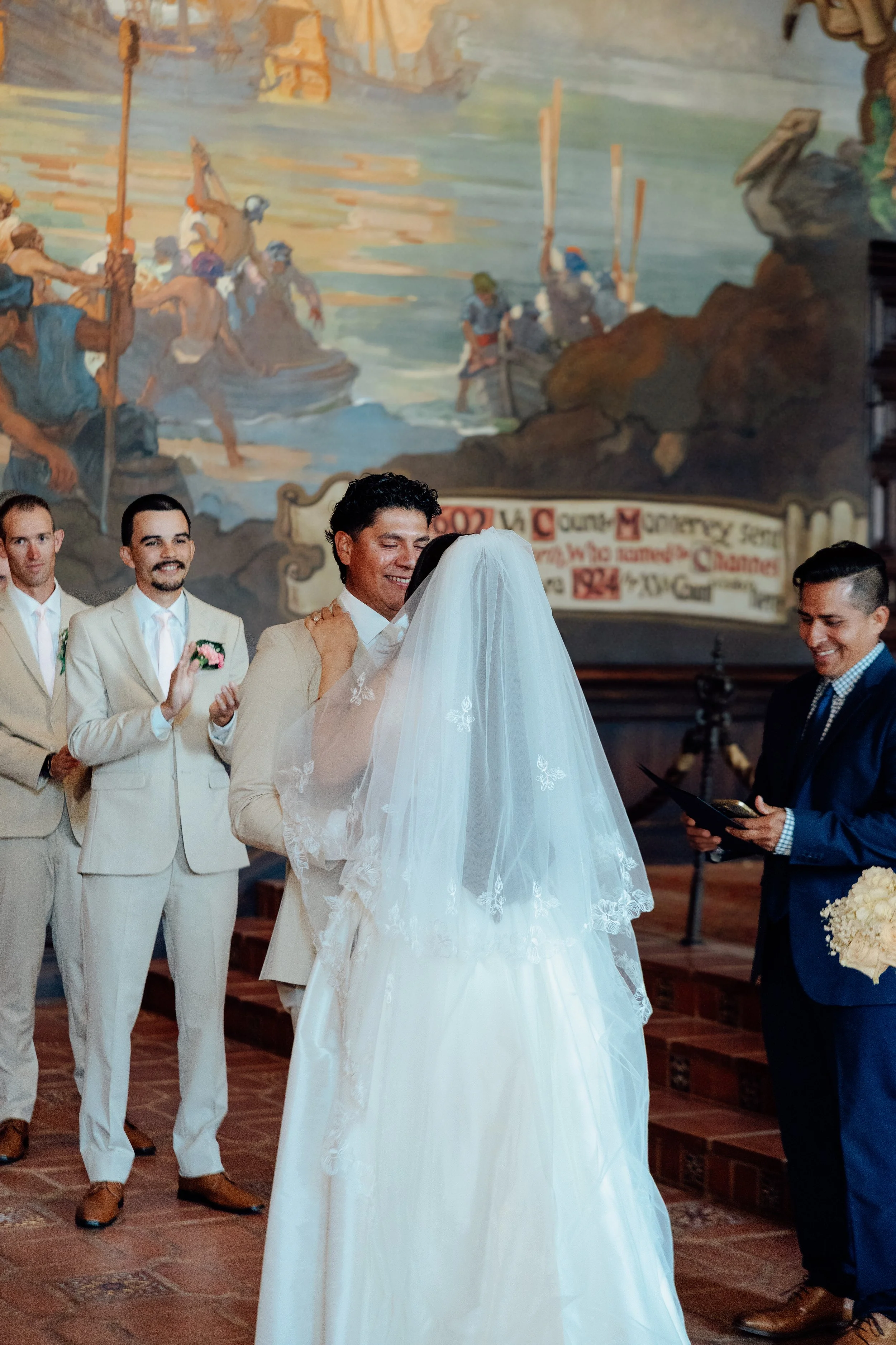 People dressed in wedding attire celebrating a wedding ceremony inside a church with a large mural painting on the wall in the background.