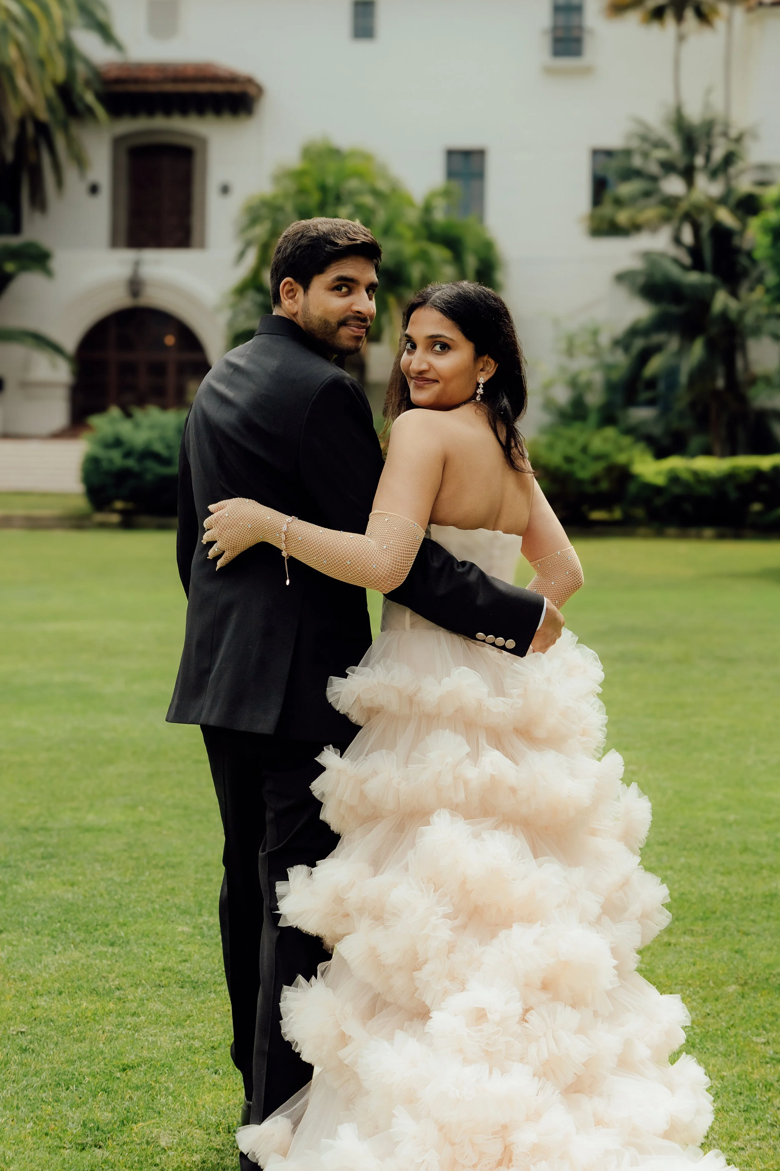 A couple dressed in wedding attire, standing outdoors on a lawn with greenery and a large white building in the background. The woman wears a strapless, tiered, peach-colored wedding gown with long, sheer gloves, and the man wears a black tuxedo. The