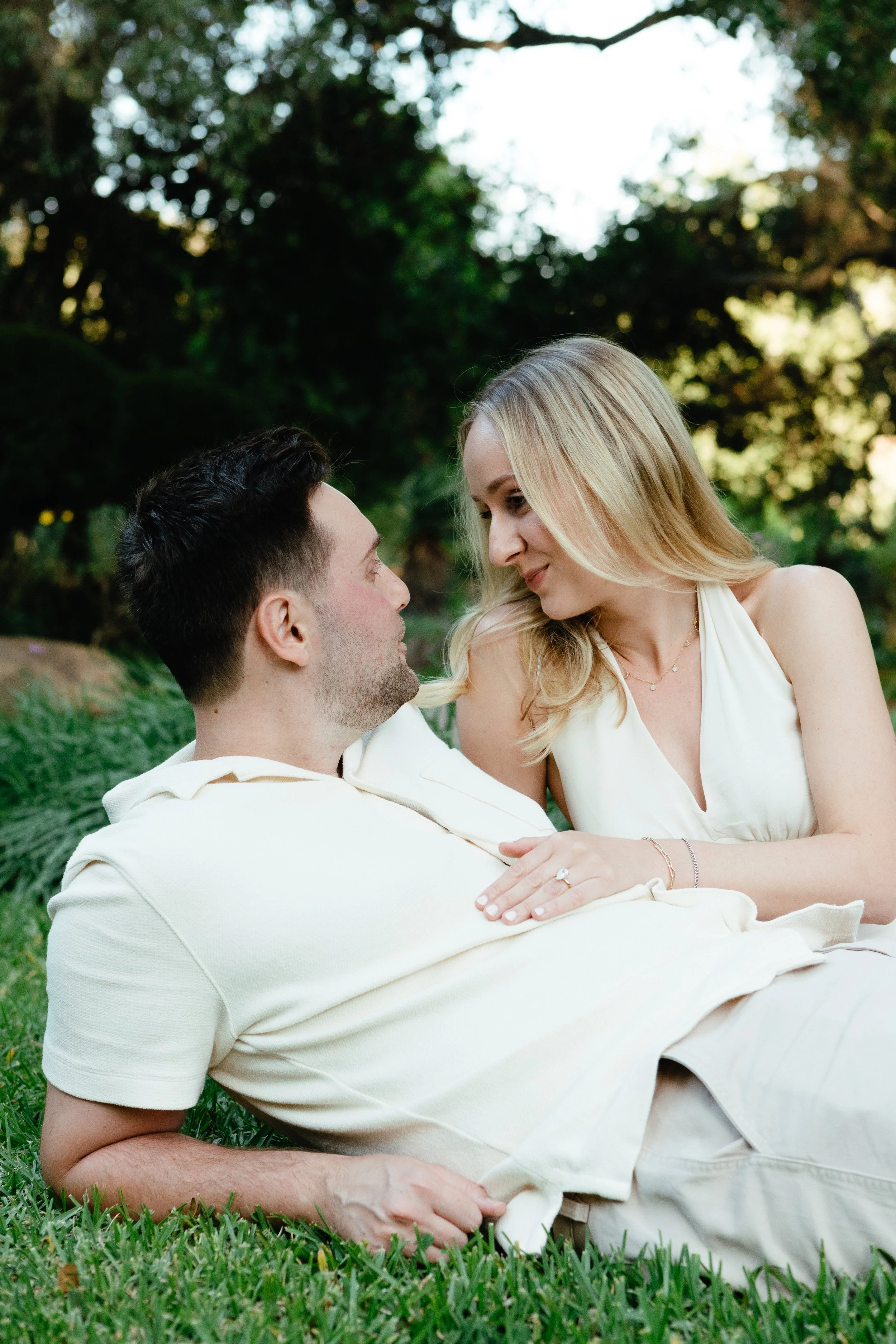 A couple sitting on the grass outdoors, embracing each other and looking into each other's eyes, with greenery in the background.
