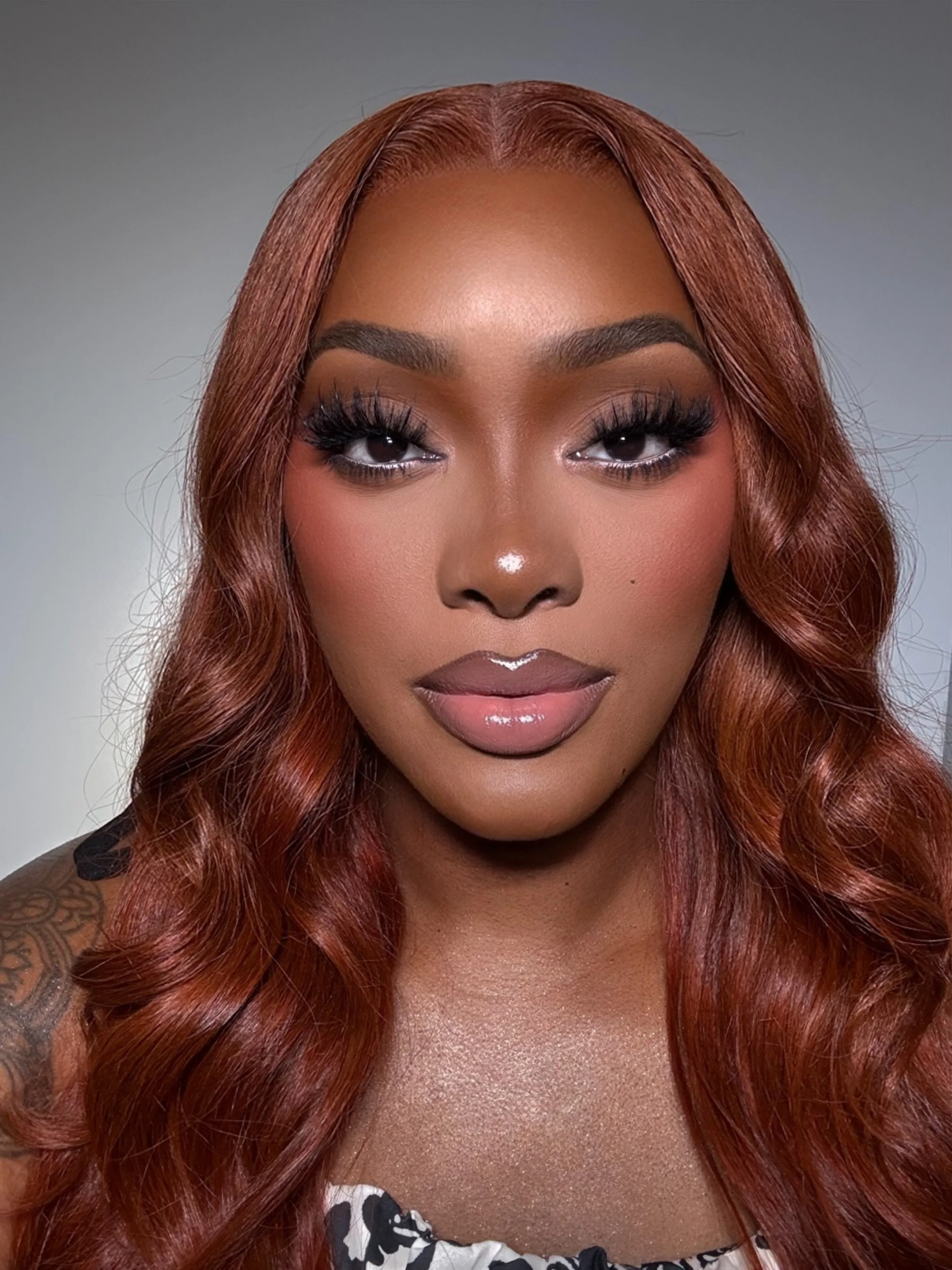 A close-up portrait of a woman with long, wavy auburn hair, makeup with bold eye makeup, and wearing a patterned top.