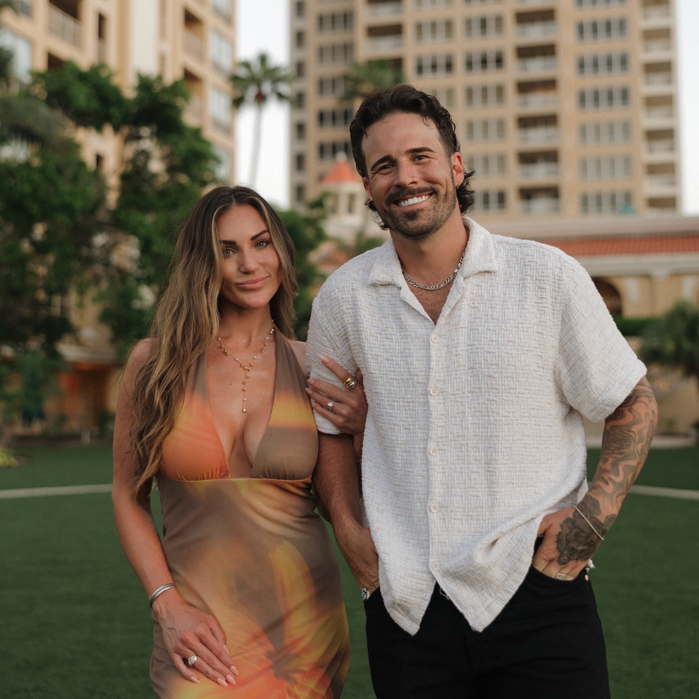 A man and woman standing outdoors in front of high-rise buildings and palm trees, smiling at the camera. The woman is wearing a dress with sunset colors and the man is wearing a white shirt with tattoos on his arm.