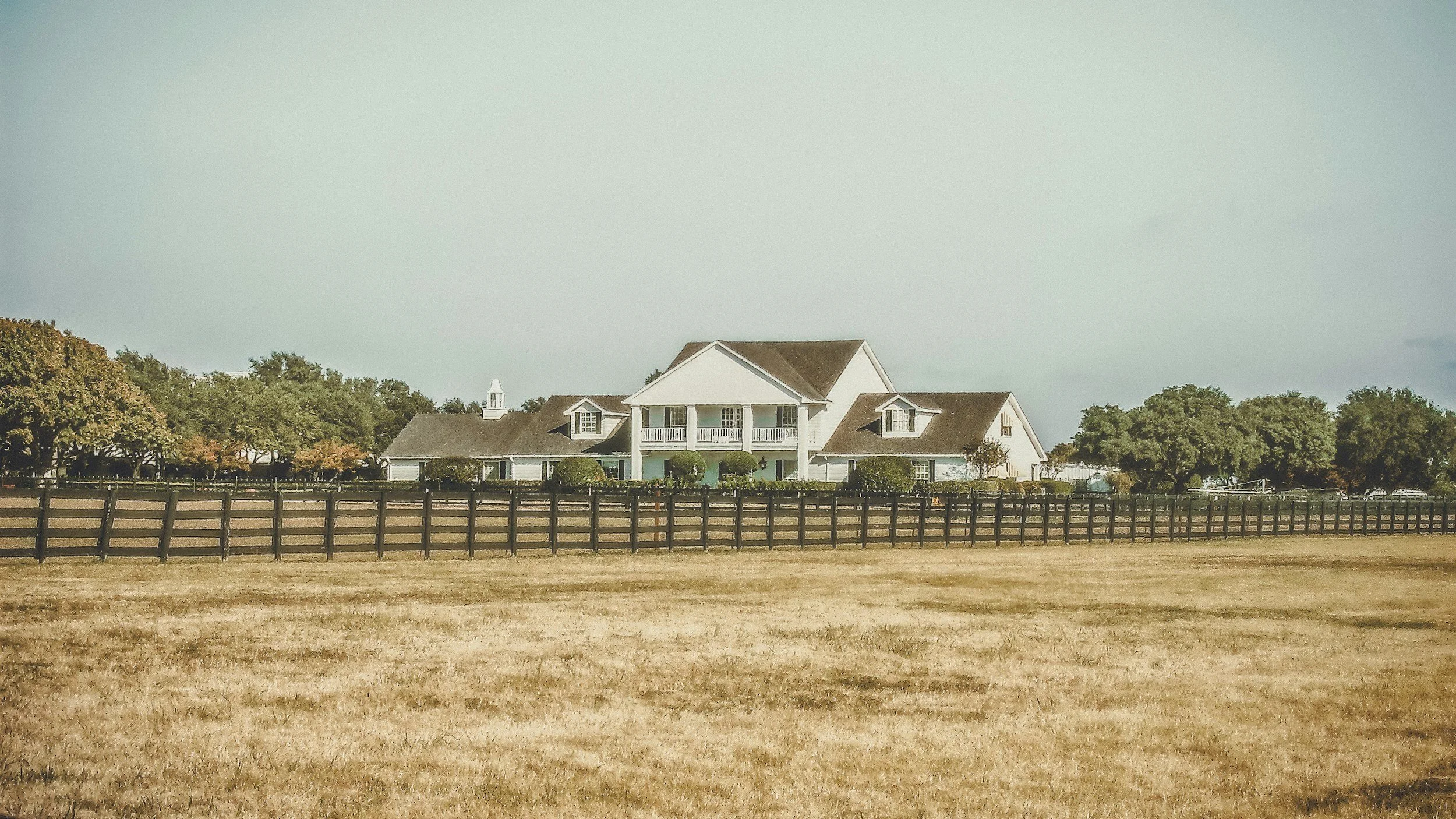 Large white house with a front porch, surrounded by trees, and enclosed by a black fence, set in a grassy field.